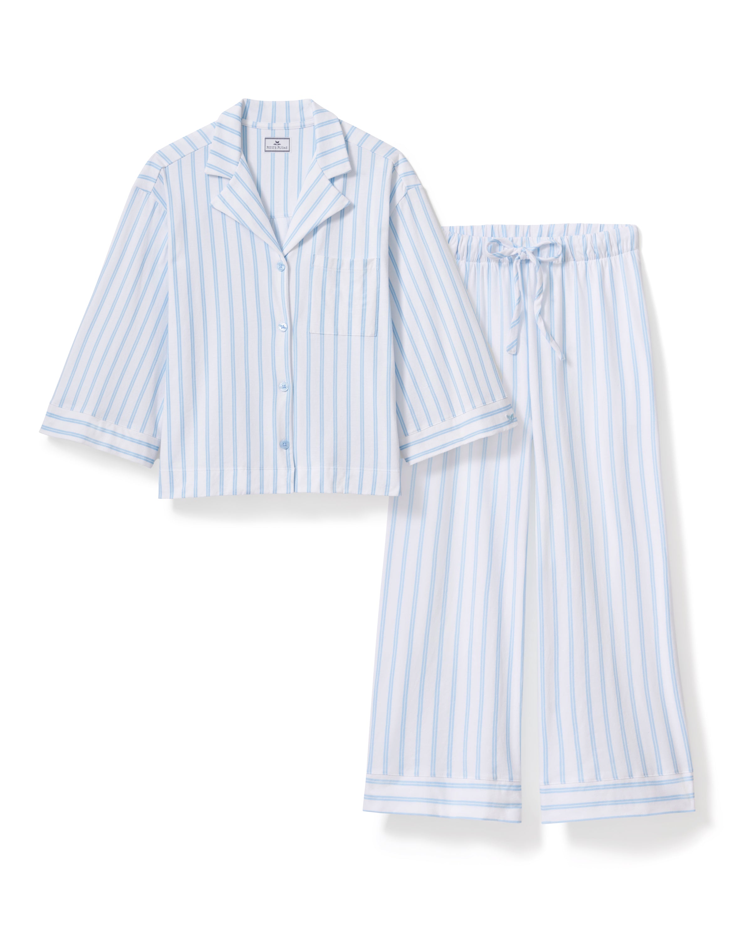 A person models the Petite Plumes Olivia Wide Leg Pima Pajama Set in Periwinkle Stripe, featuring a soft, luxurious white and light blue striped button-up long-sleeve shirt with matching wide-leg pants against a plain background that enhances its elegant simplicity.