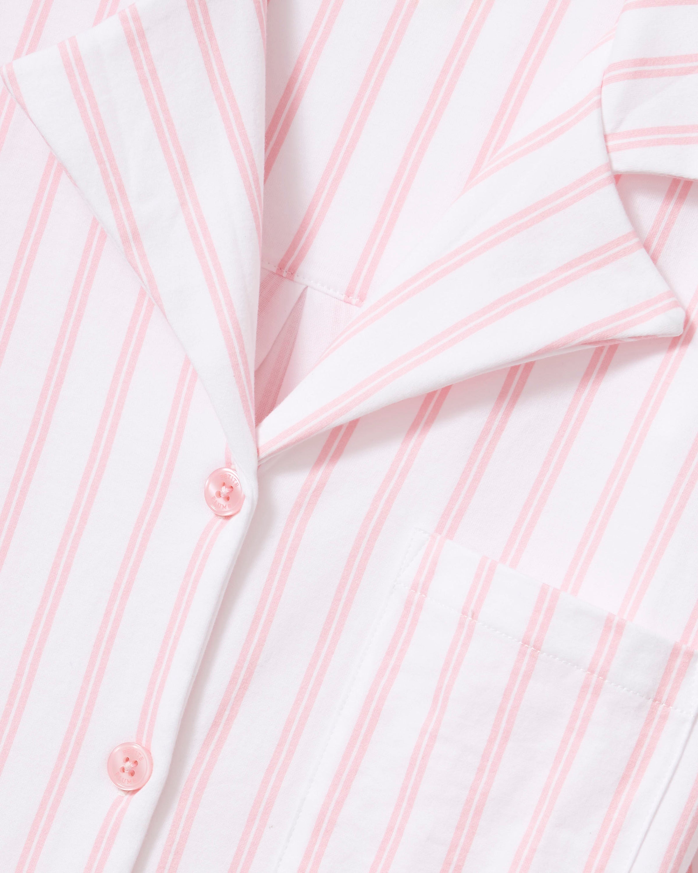 A person is seen in The Olivia Wide Leg Pima Pajama Set in Pink Stripe by Petite Plume, featuring a button-up top with a collar and long sleeves, and wide-leg pants made of luxurious Peruvian Pima cotton. The backdrop is plain and light-colored.