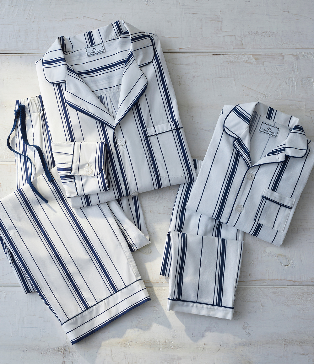 Two sets of white pajamas with blue vertical stripes are neatly folded on a light wooden surface. The sets include long-sleeve tops with collars, button fronts, and matching pants.