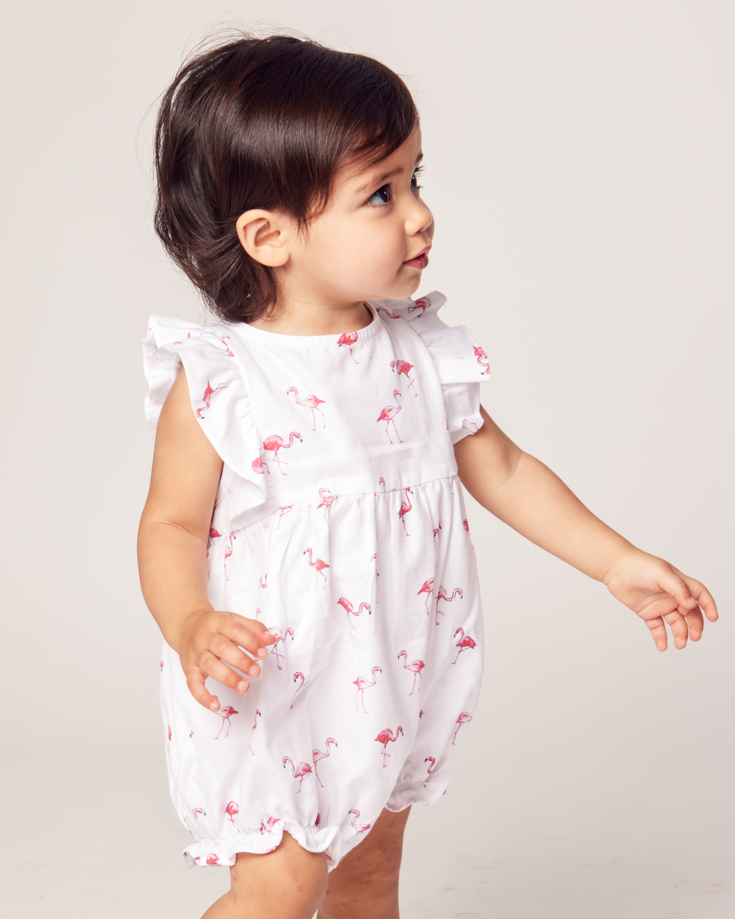 A toddler wearing Petite Plumes Babys Twill Ruffled Romper in Flamingos, featuring luxury cotton with pink flamingo prints and ruffled sleeves, gazes to the side. The tropical pajama vibe pops against the plain light-colored background.
