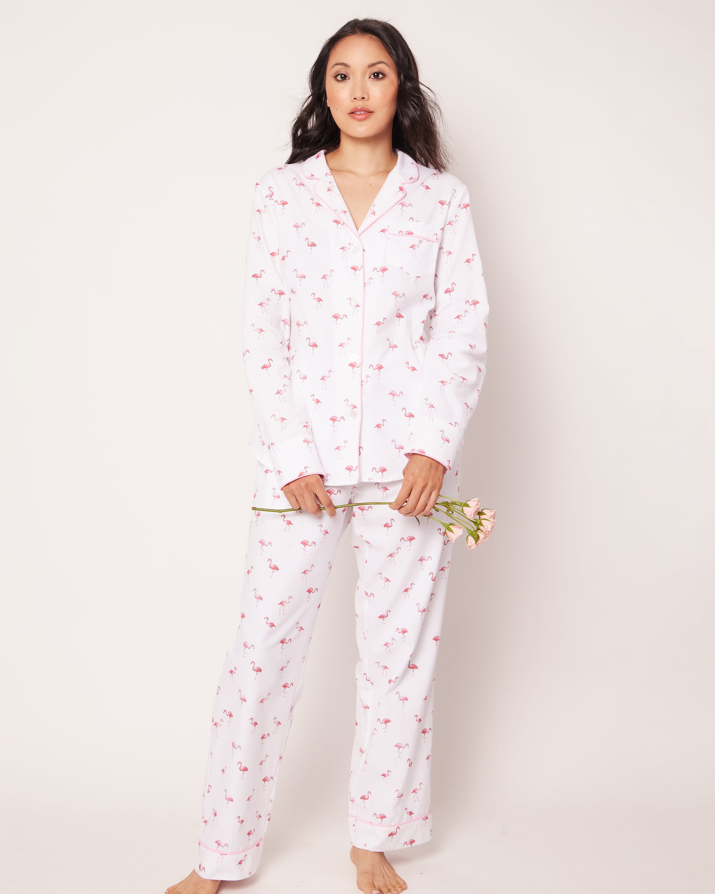 A person in Petite Plumes Womens Twill Pajama Set in Flamingos, featuring small pink floral patterns, holds a pink flower against a plain, light-colored background, evoking tropical twist elegance.
