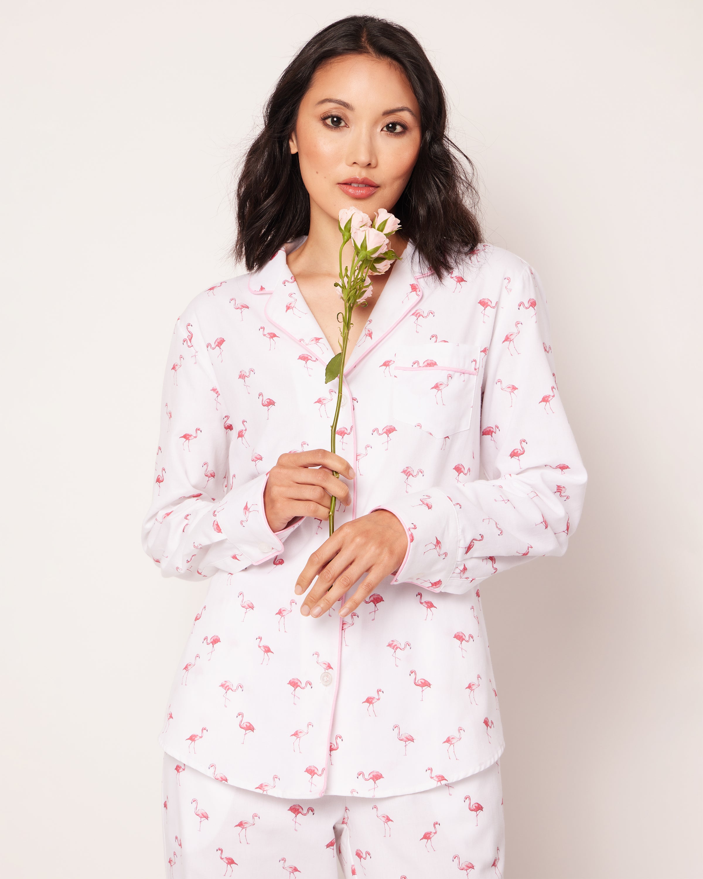 A woman in Petite Plume’s Womens Twill Pajama Set in Flamingos holds a pink flower against a plain background, glancing at the camera with a slight smile, adding tropical charm to her serene demeanor.