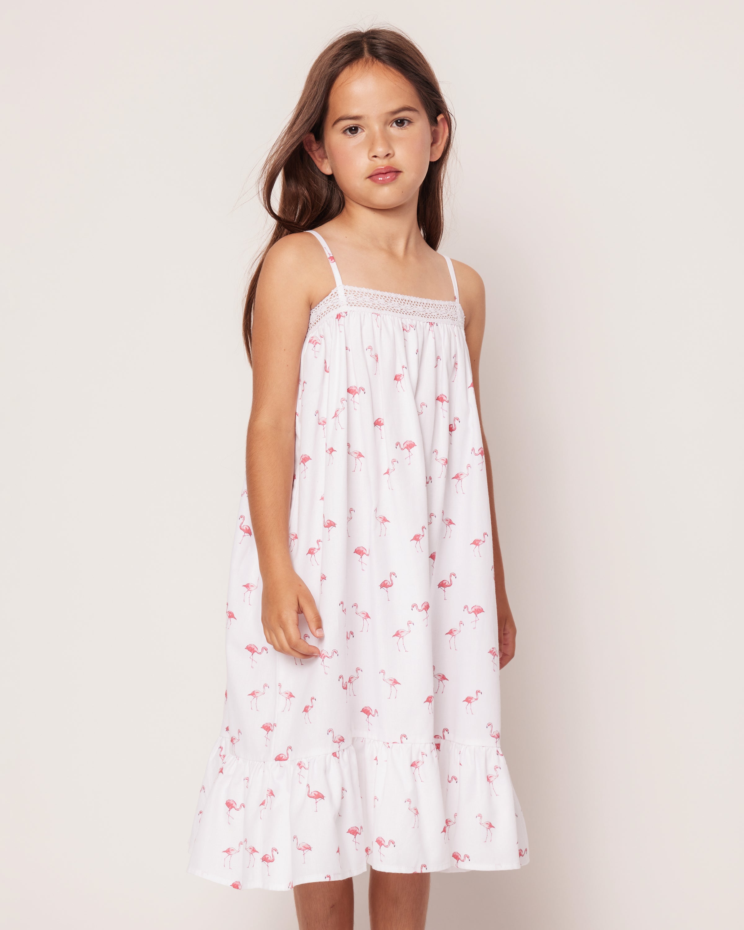 A young girl with long brown hair wears Petite Plumes Girls Twill Lily Nightgown in Flamingos, featuring thin straps and a ruffled hem. She stands against a plain off-white background with a neutral expression, the small pink flamingo pattern adding a tropical twist.