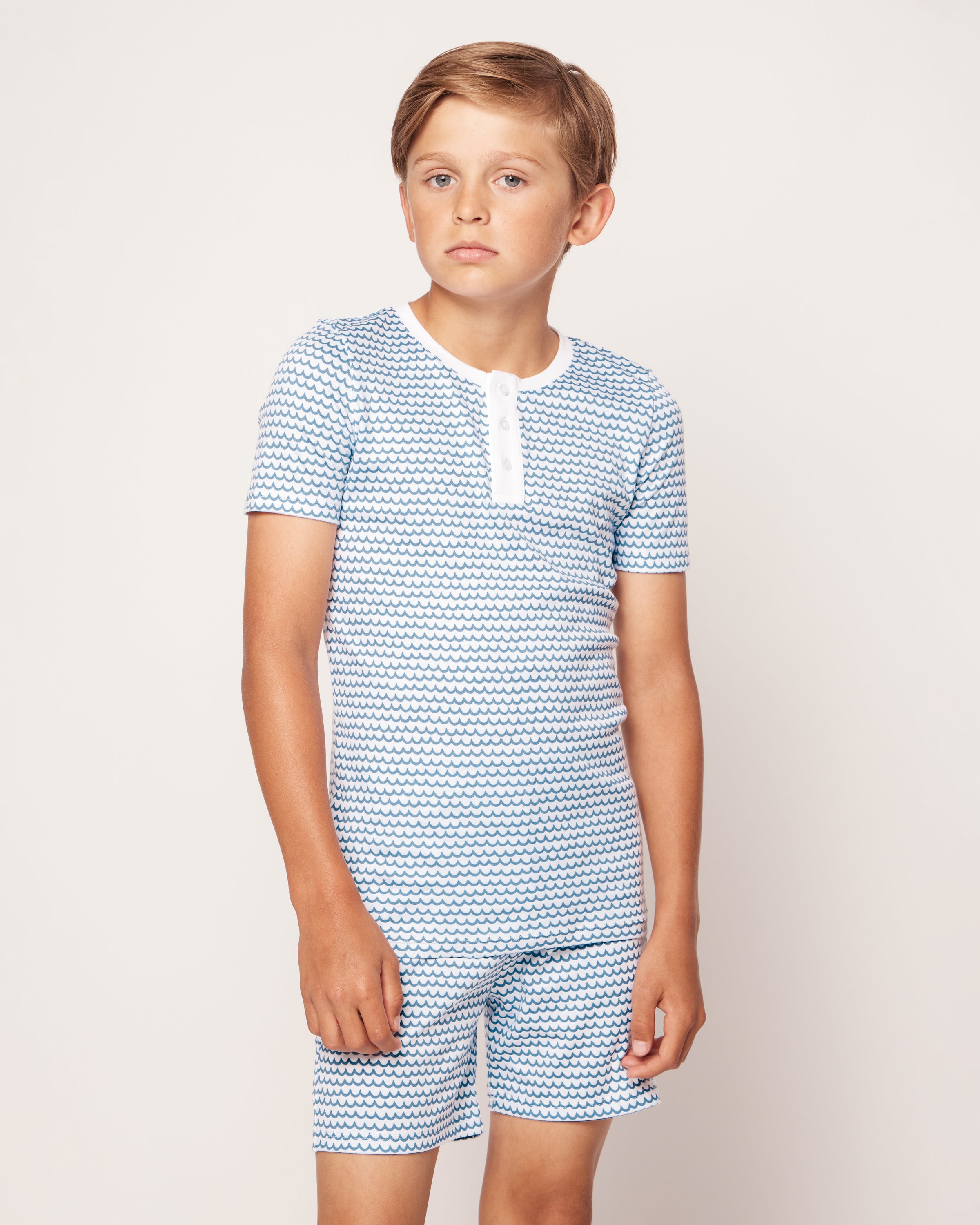 A young boy with short light brown hair stands against a plain background, wearing the Kids Pima Snug Fit Pajama Short Set in La Mer by Petite Plume. His sleepwear ensemble includes a blue and white patterned top with matching shorts, and he has a neutral expression.