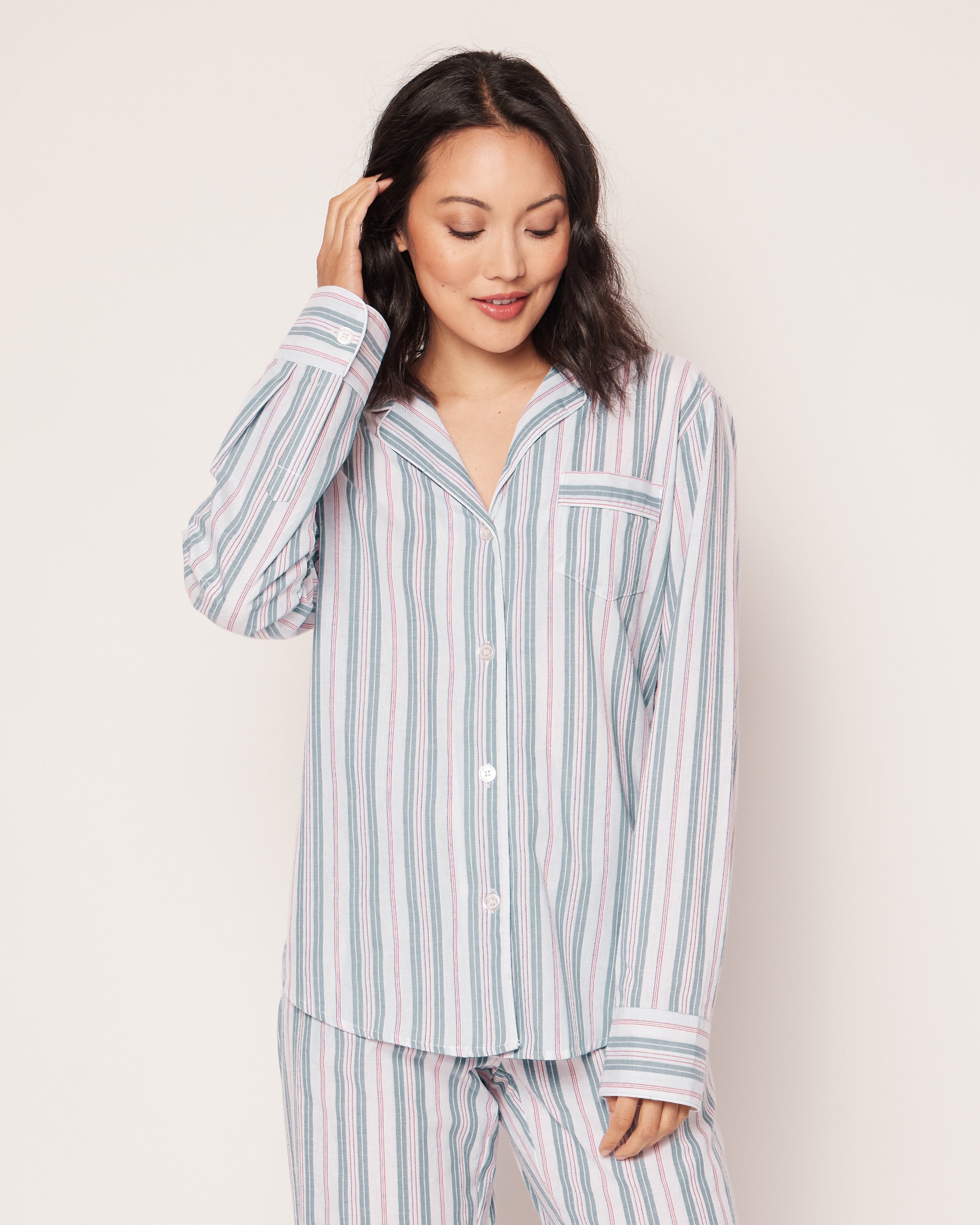 A woman gently smiles, touching her hair in a Petite Plume Womens Twill Pajama Set in Vintage French Stripes, enjoying luxury sleepwear with a button-up top and matching pants in white, blue, and pink. The plain light background enhances her serene appearance.