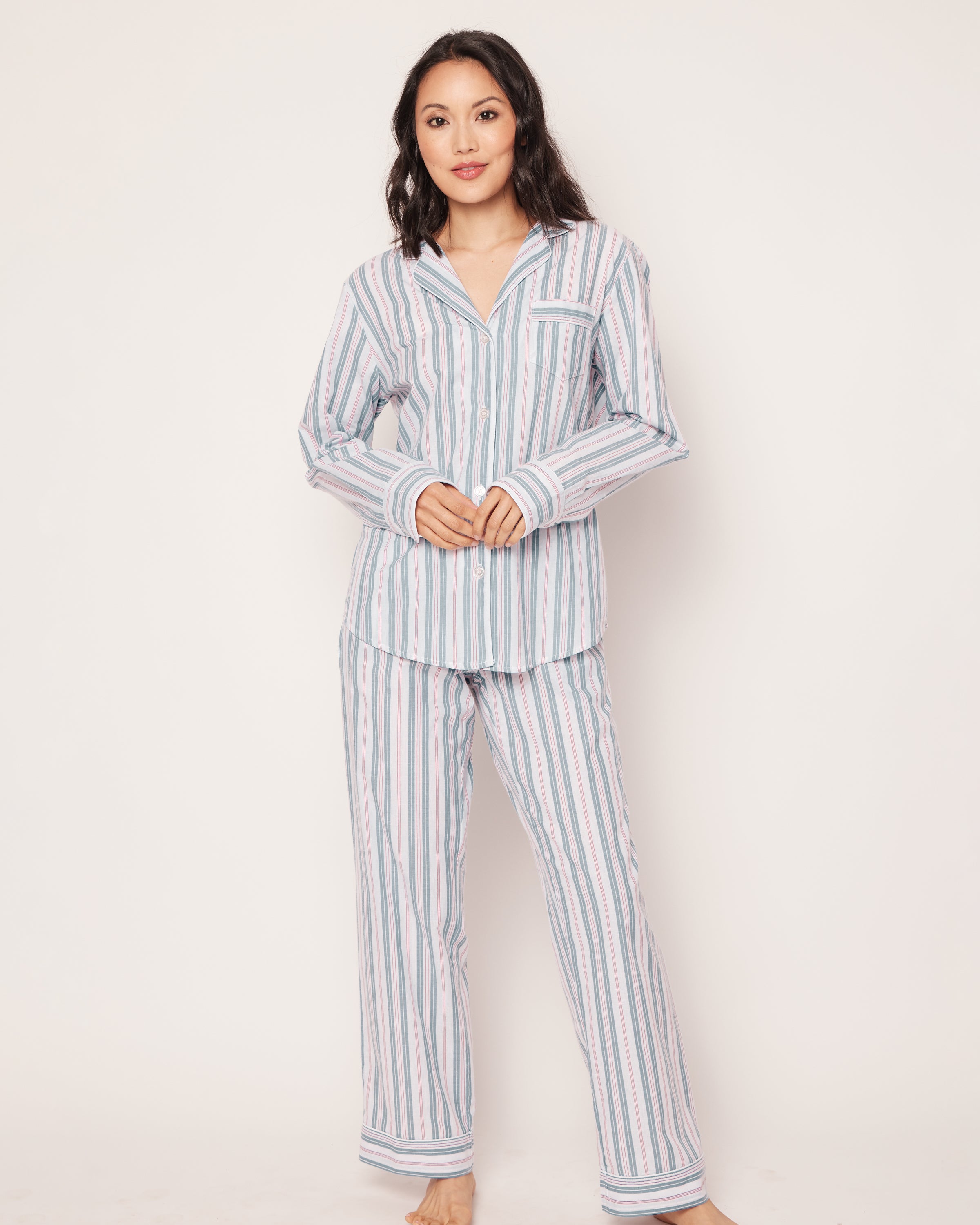 Dressed in a Womens Twill Pajama Set in Vintage French Stripes by Petite Plume, a person with long, dark hair stands barefoot against a plain background. The light-colored pajamas feature vertical stripes and include a button-up shirt with a collar and pocket. They gaze directly at the camera.