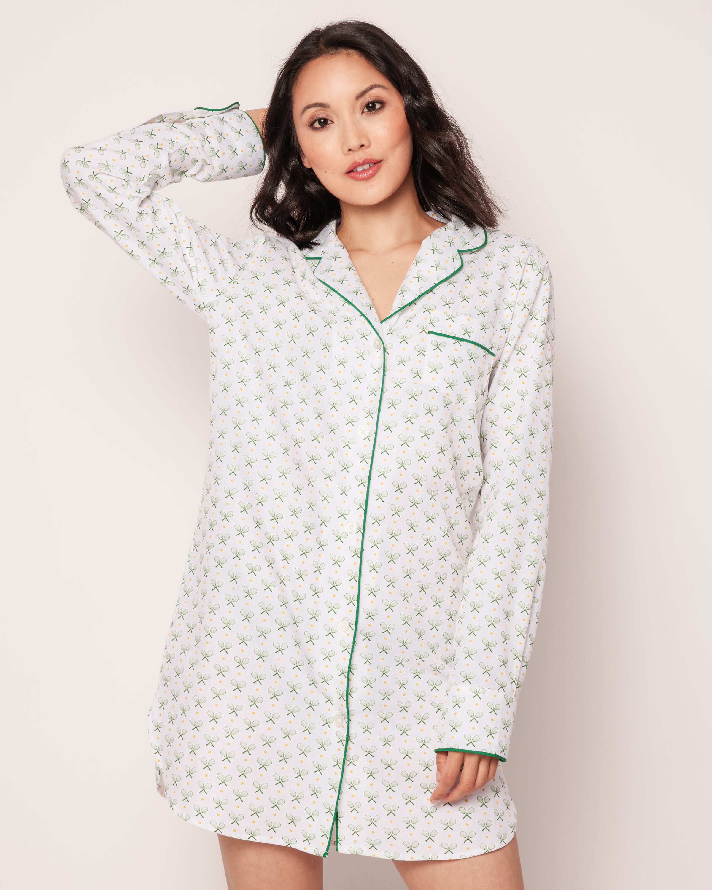 A woman in Petite Plumes Womens Twill Nightshirt in Match Point, featuring a subtle pattern with green piping, poses with one hand behind her head against a plain background.
