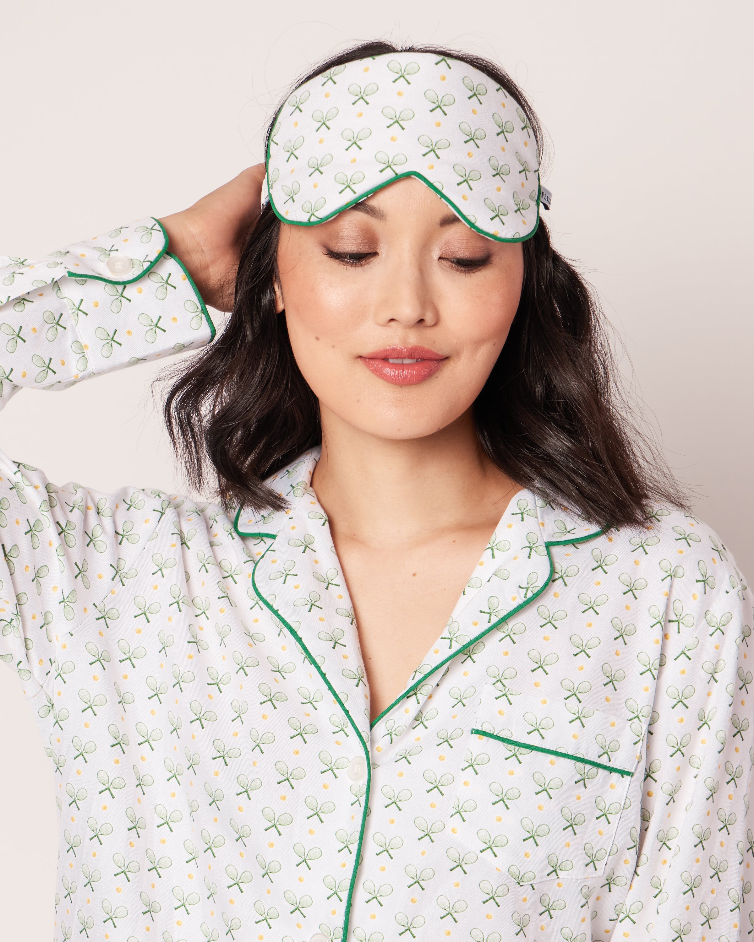A person wearing tennis print pajamas adjusts a Petite Plume Adults Sleep Mask in Match Point on their forehead. The white mask with green detailing complements the plain light-colored background, and the individuals long dark hair completes this cozy yet stylish ensemble.