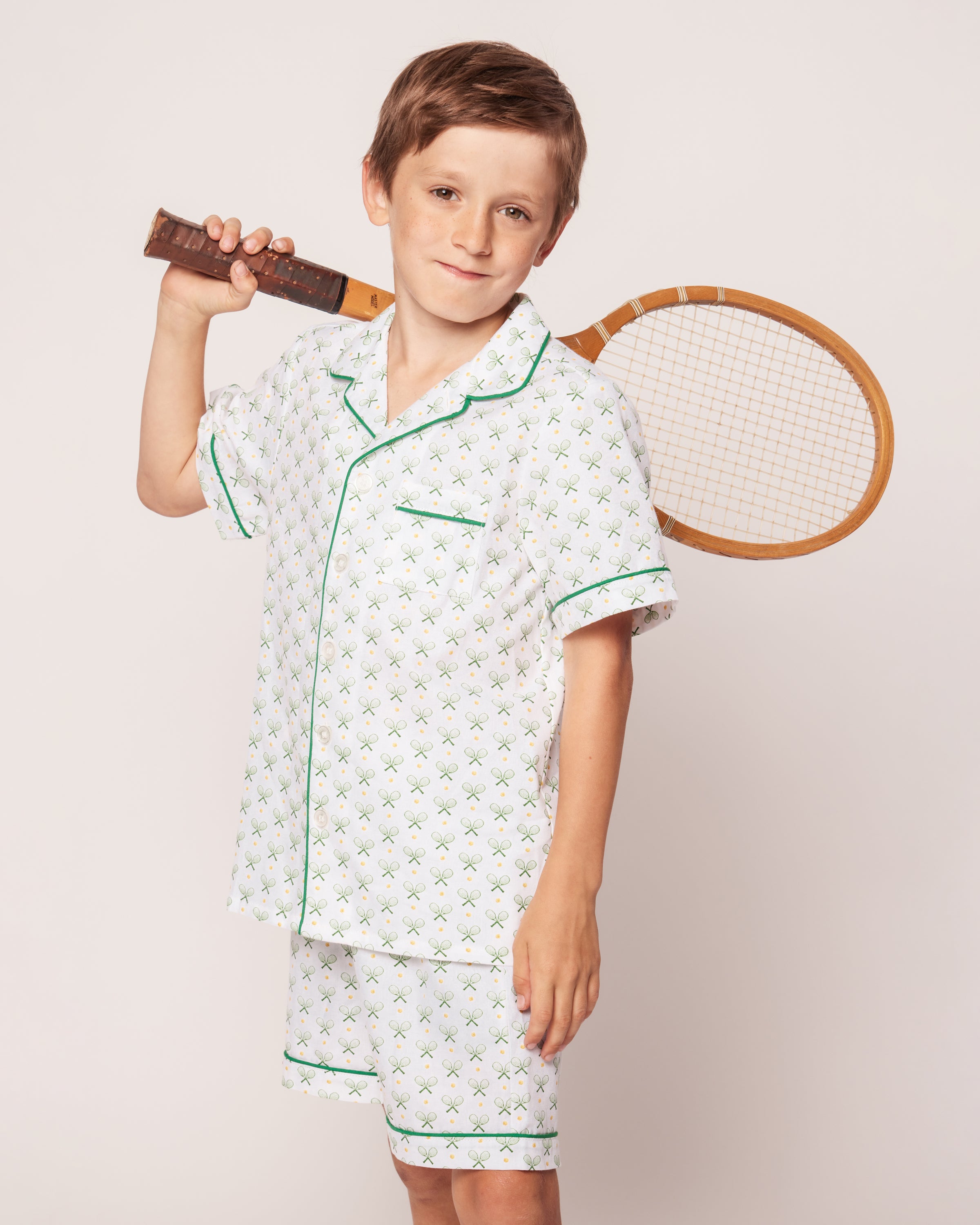 A young boy, holding a vintage wooden tennis racket over his shoulder, is dressed in Petite Plumes Kids Twill Pajama Short Set in Match Point. The luxury sleepwear, featuring a green and blue pattern, stands out against the white background, offering comfort and style for restful nights.