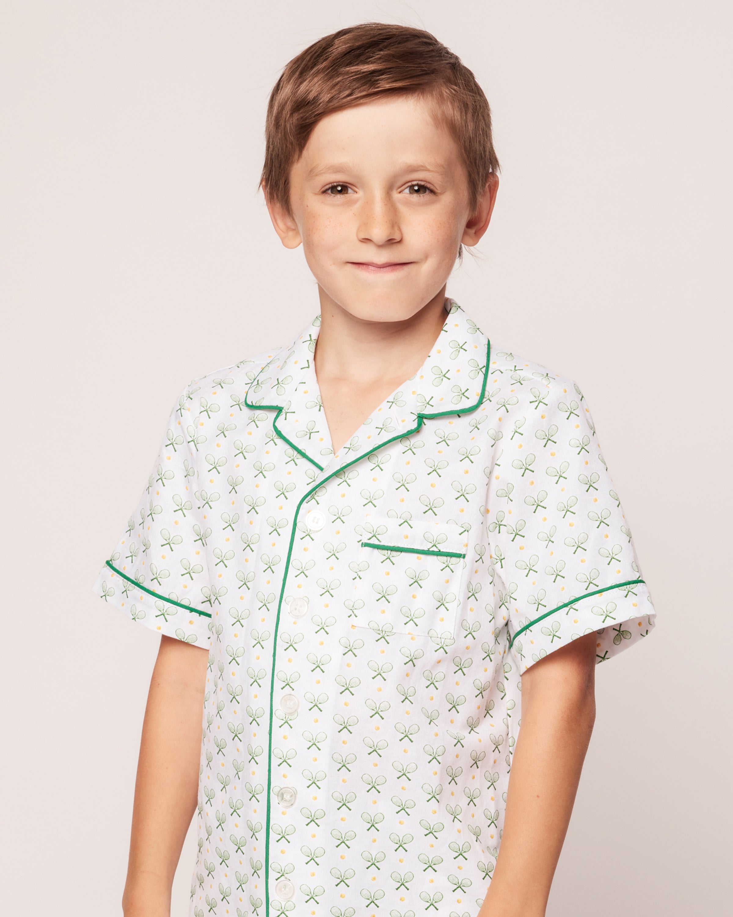 A young boy, effortlessly stylish, smiles at the camera in a Petite Plume Kids Twill Pajama Short Set in Match Point. The set features a white base with a classic tennis print and green piping on the collar, sleeves, and pocket against a simple background for added charm.