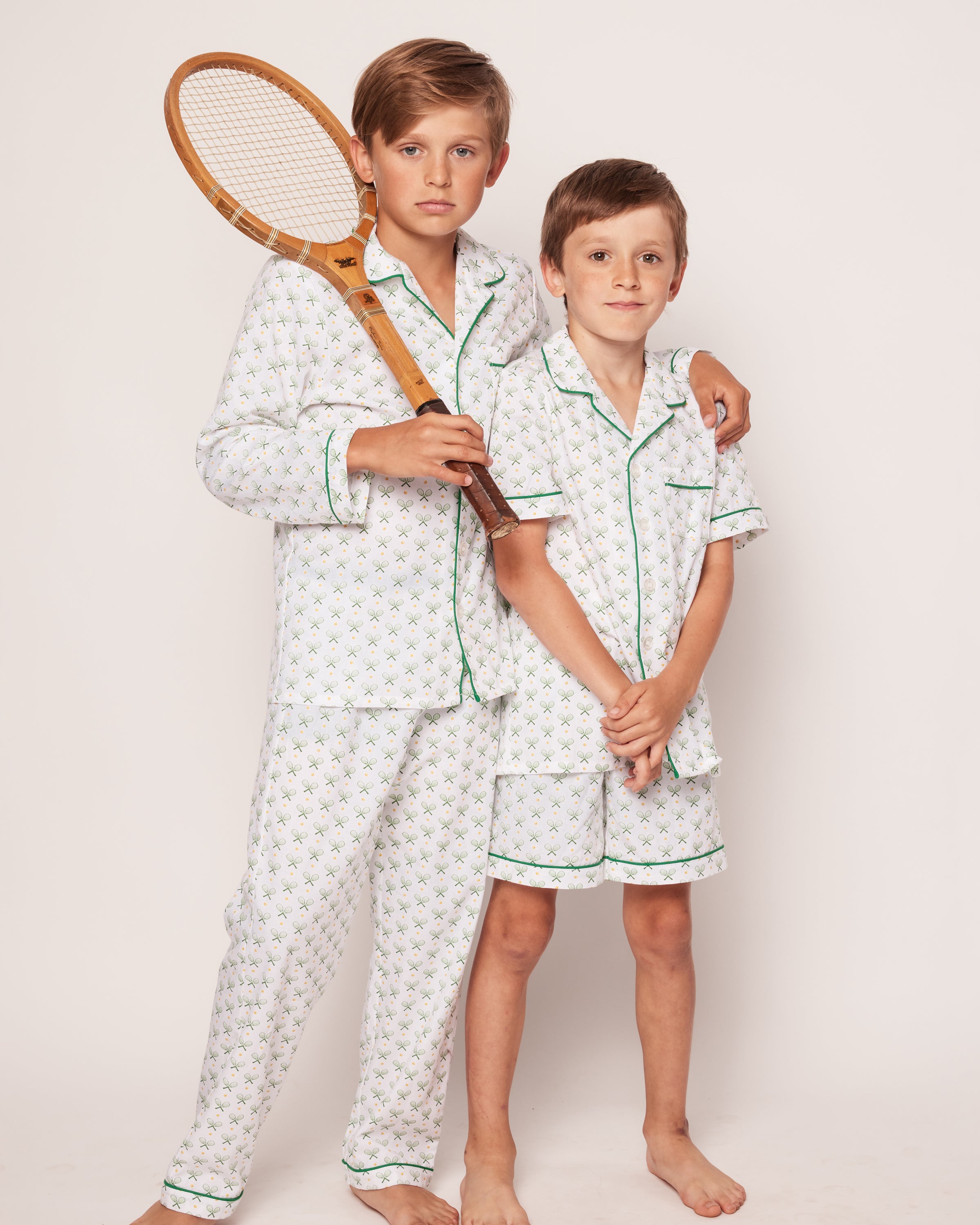 Two young boys, barefoot against a white background, wear matching Kids Twill Pajama Short Sets in Match Point by Petite Plume. The boy on the left holds a vintage wooden tennis racket and gently wraps his arm around the younger boy, both exuding elegance in luxurious sleepwear.