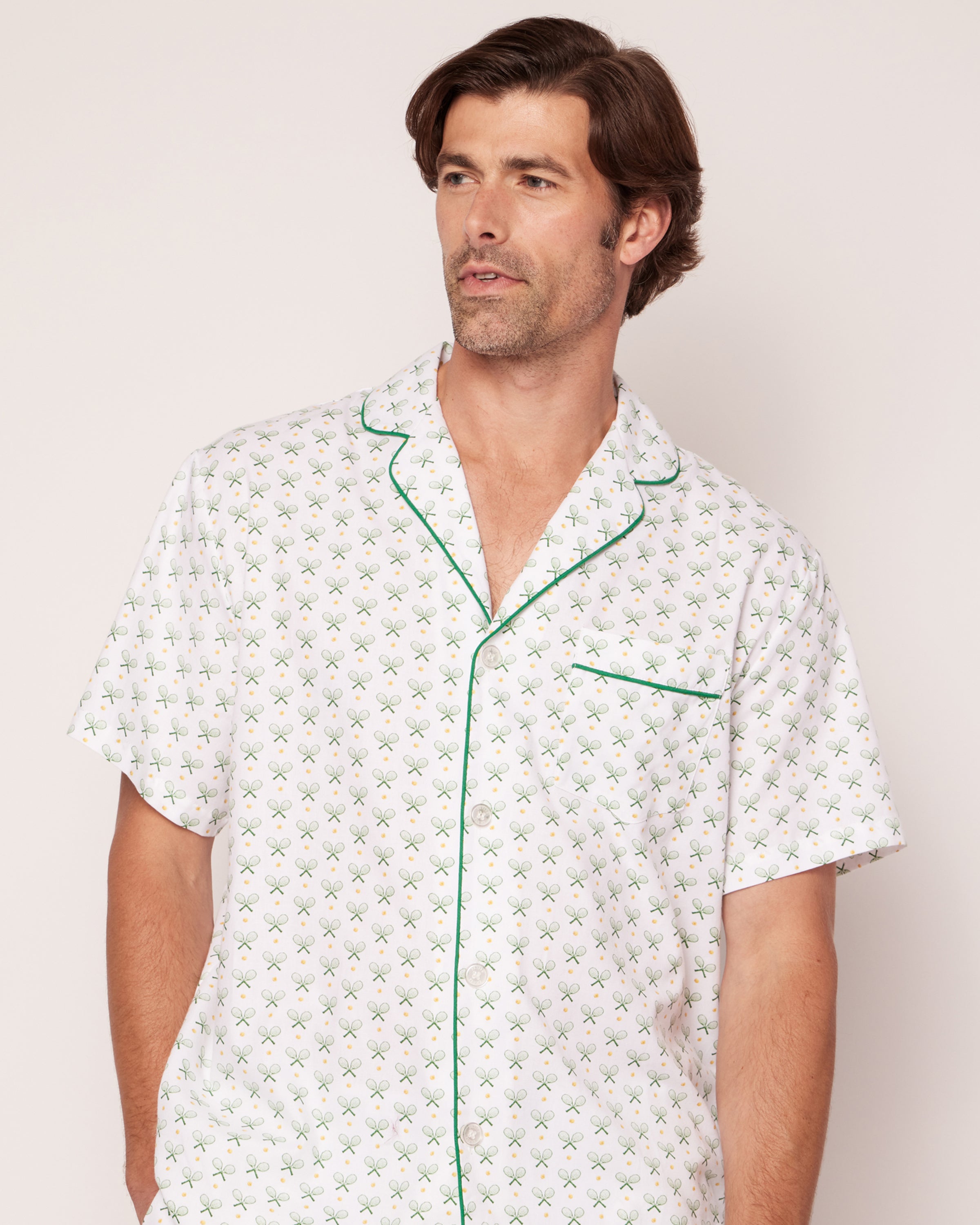 A man models the Match Point pajama top from Petite Plume—a white 100% cotton piece with a green geometric design, reminiscent of tennis print pajamas. It includes green piping and a left-side pocket. With brown hair, he gazes sideways against a plain background, exuding luxurious sleepwear style.