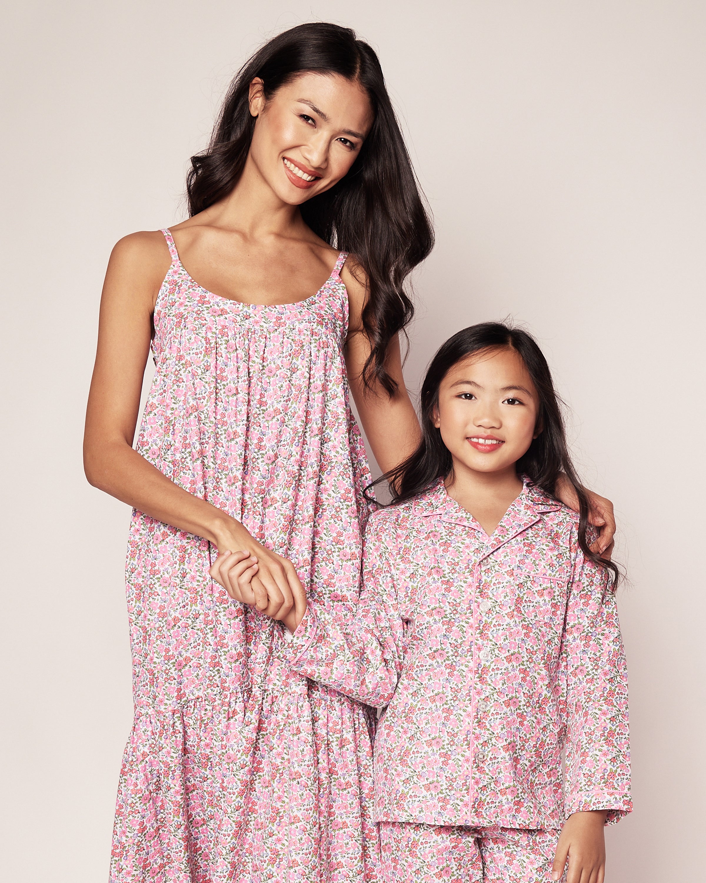 A woman and a young girl smile in matching luxury sleepwear from Petite Plume, wearing the Womens Twill Chloe Nightgown in Fleurs de Rose and pajamas. They stand against a neutral background, holding hands affectionately.