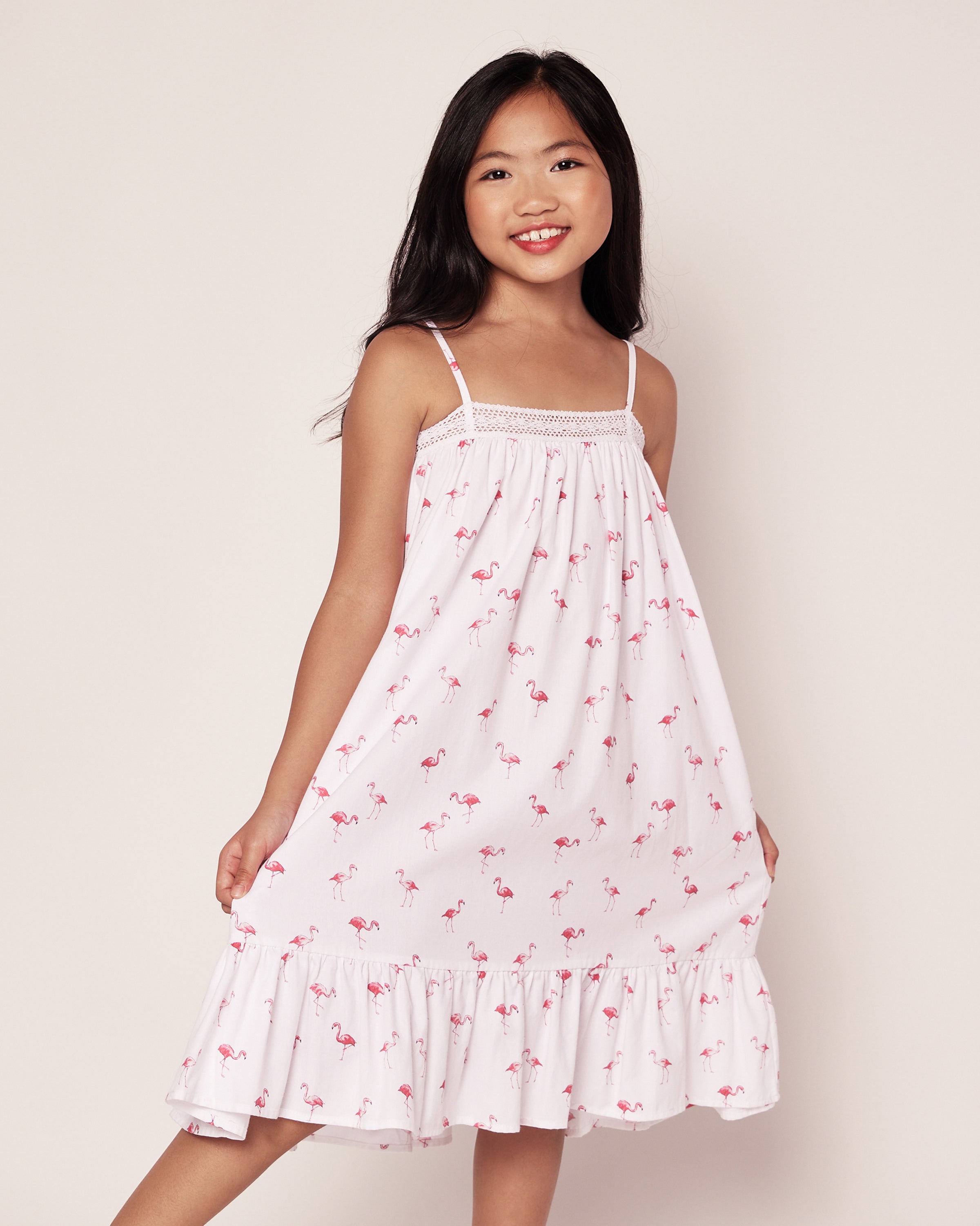 A young girl in the Petite Plume Girls Twill Lily Nightgown featuring pink flamingos smiles against a light background, showcasing its sleeveless design and ruffled hem—a perfect tropical twist.