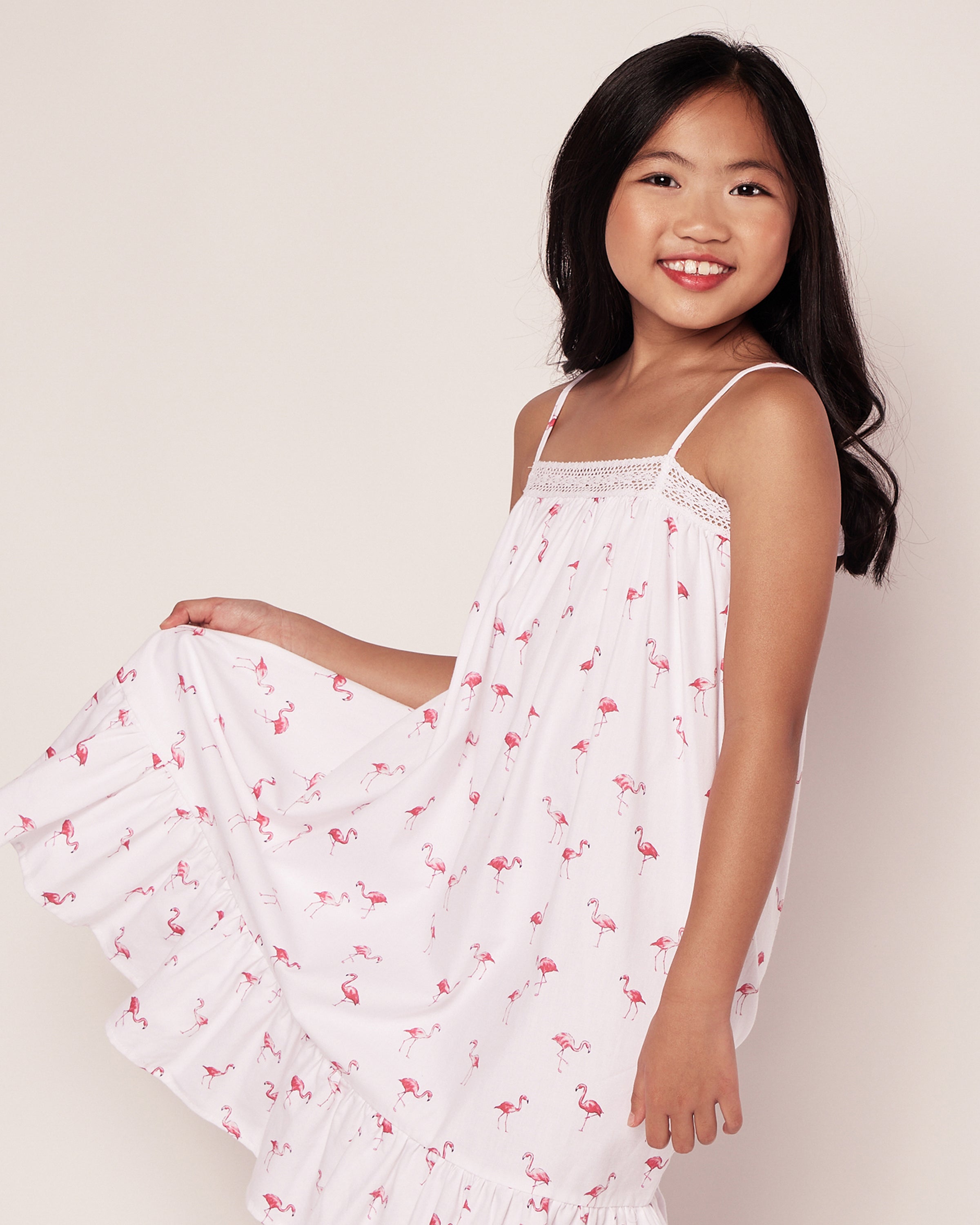 A young girl, with long dark hair, smiles while holding out the skirt of her Girls Twill Lily Nightgown in Flamingos by Petite Plume against a light background.