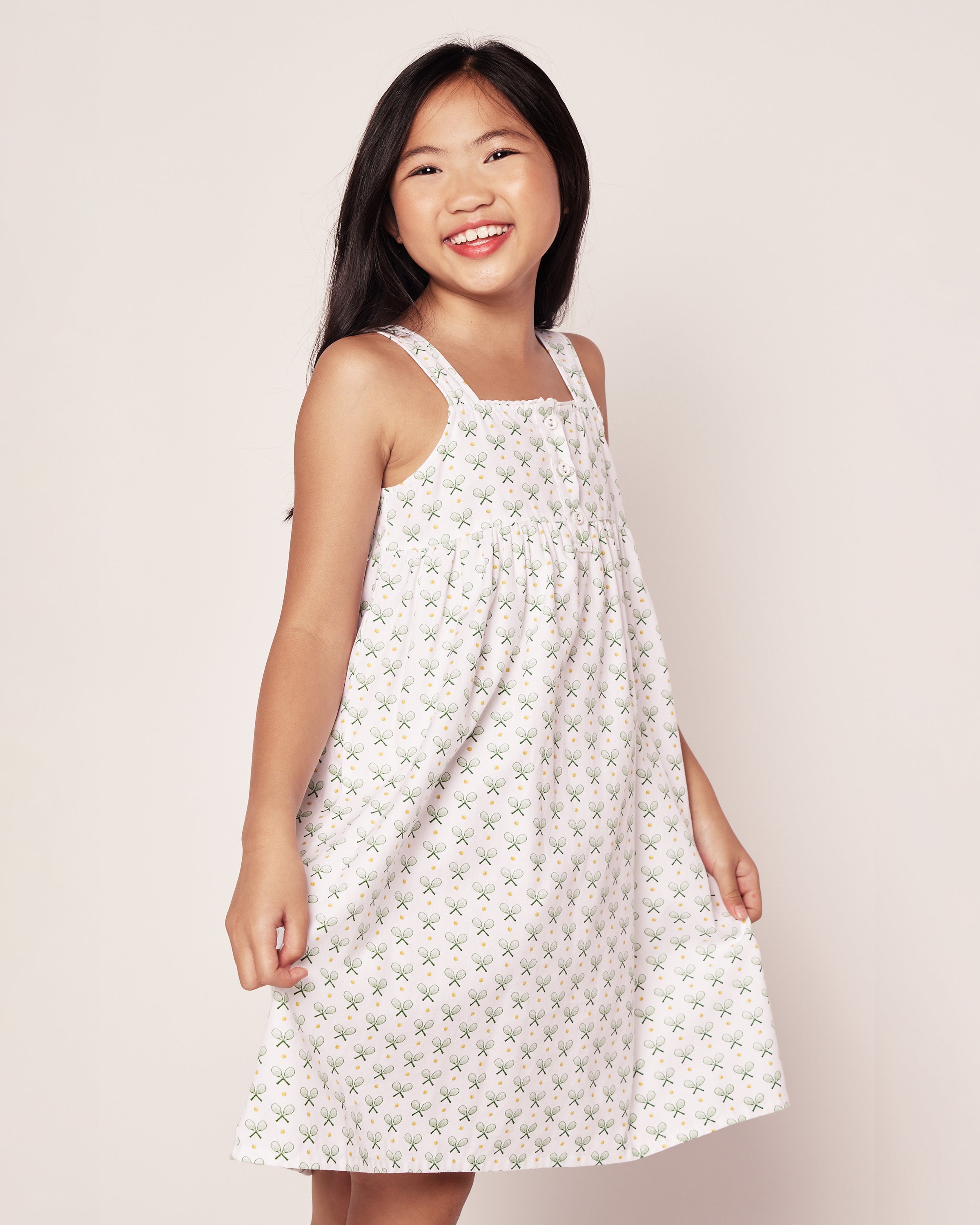 A young girl beams in Petite Plumes Girls Twill Charlotte Nightgown in Match Point. She stands against a light backdrop, her long hair flowing as she gazes sideways, exuding comfort and style with its small floral designs reminiscent of tennis print pajamas.