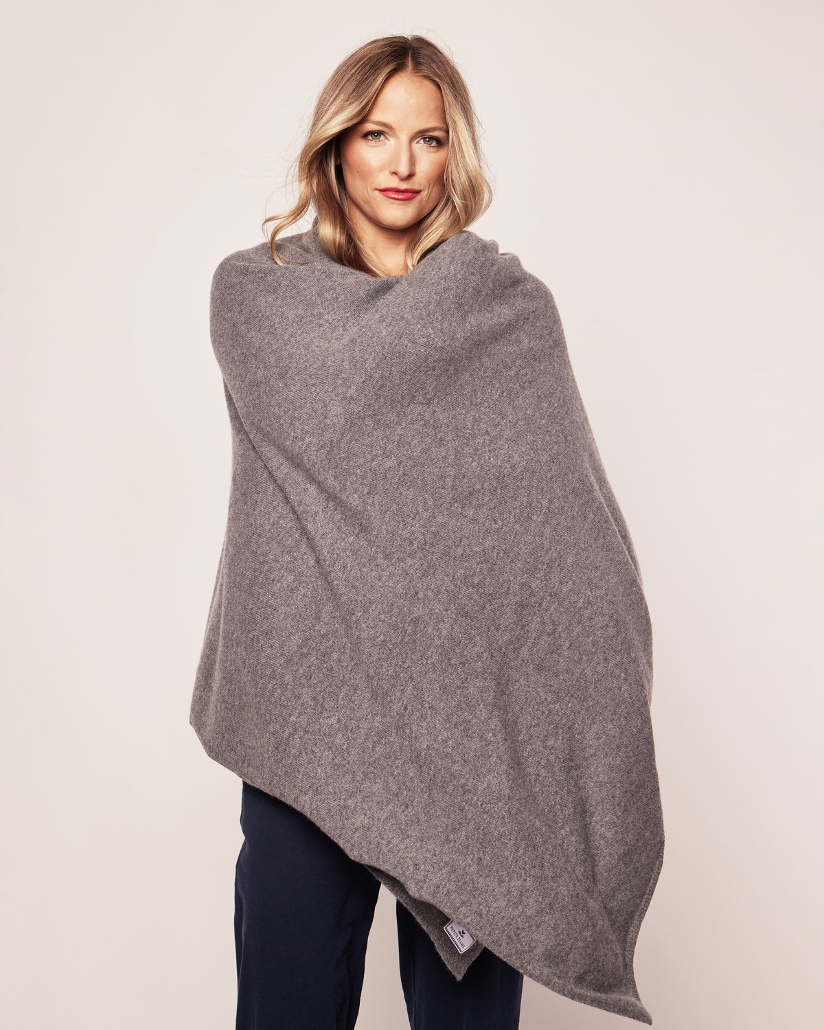 A woman with long blonde hair stands against a plain background, wrapped in Petite Plumes Womens Cashmere Wrap in Dark Grey. Wearing dark pants, she gazes directly at the camera with a neutral expression that exudes warmth and sophistication.