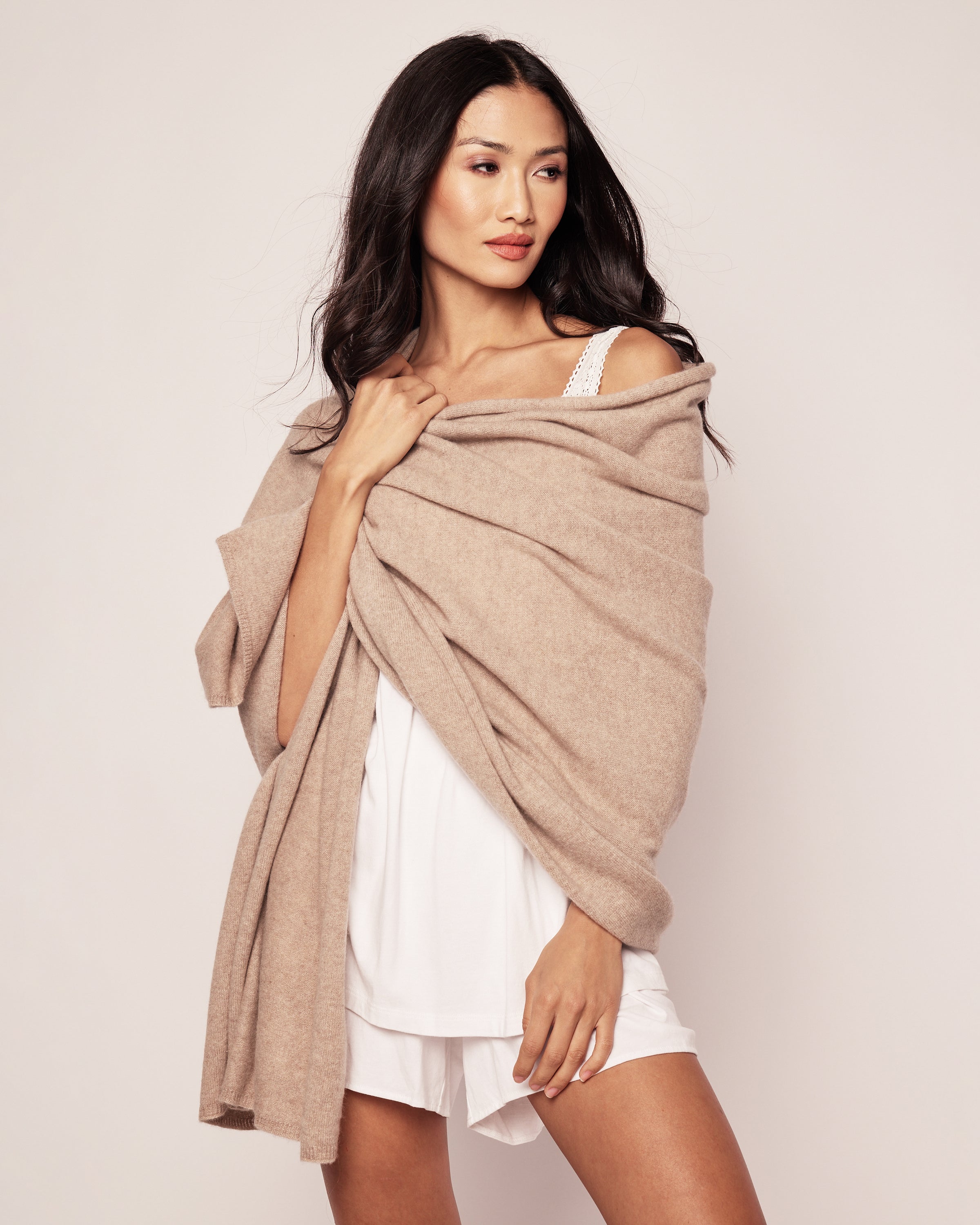 Draped in a Petite Plume Womens Cashmere Wrap in Beige over her white outfit, she exudes elegance. Her long, dark hair flows gracefully as she glances to the side with a subtle smile. The plain, light-colored background accentuates her relaxed and cozy look.