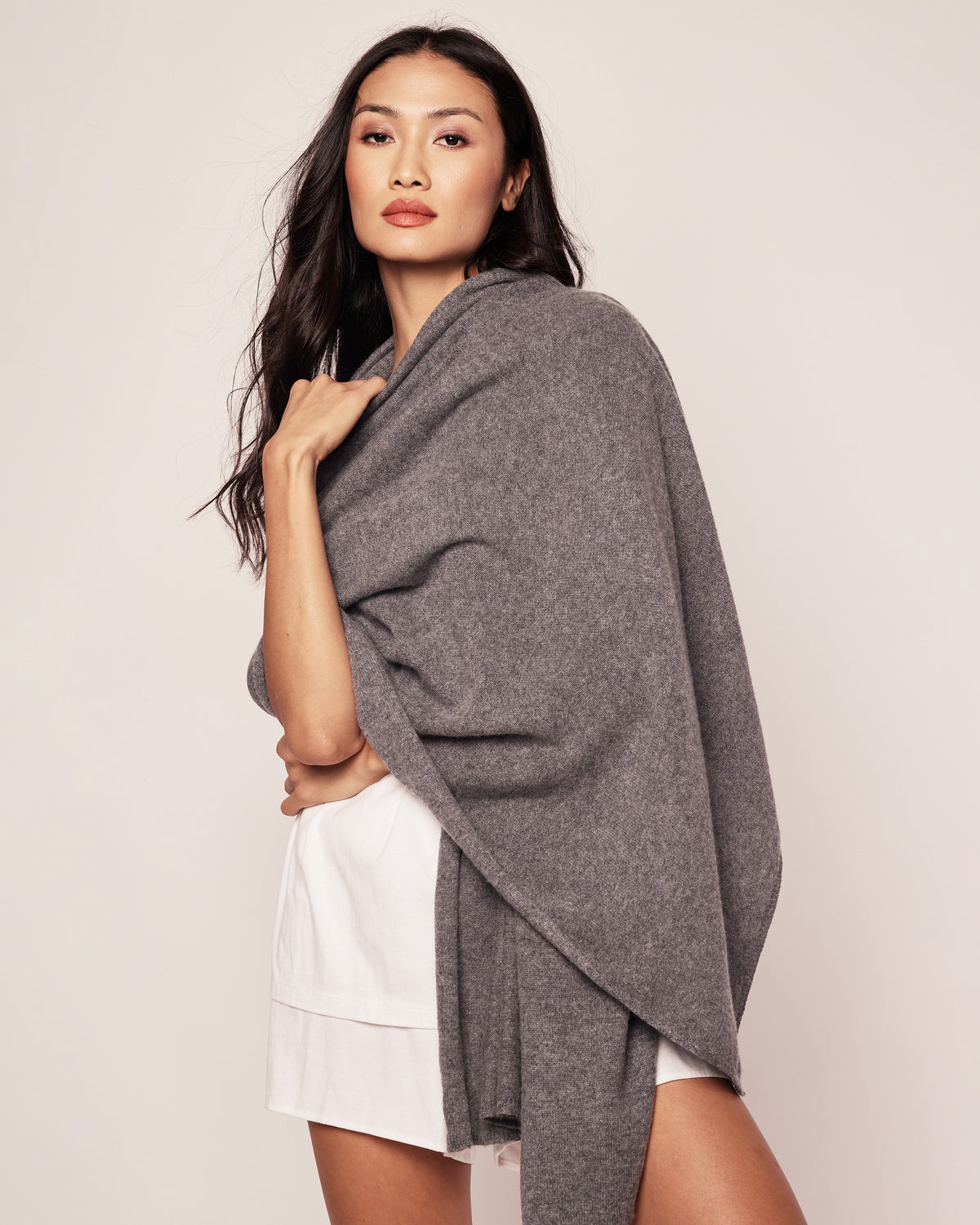 A woman with long dark hair is wrapped in Petite Plumes Womens Cashmere Wrap in Dark Grey, exuding warmth and elegance. She confidently poses in a white outfit against an off-white background.