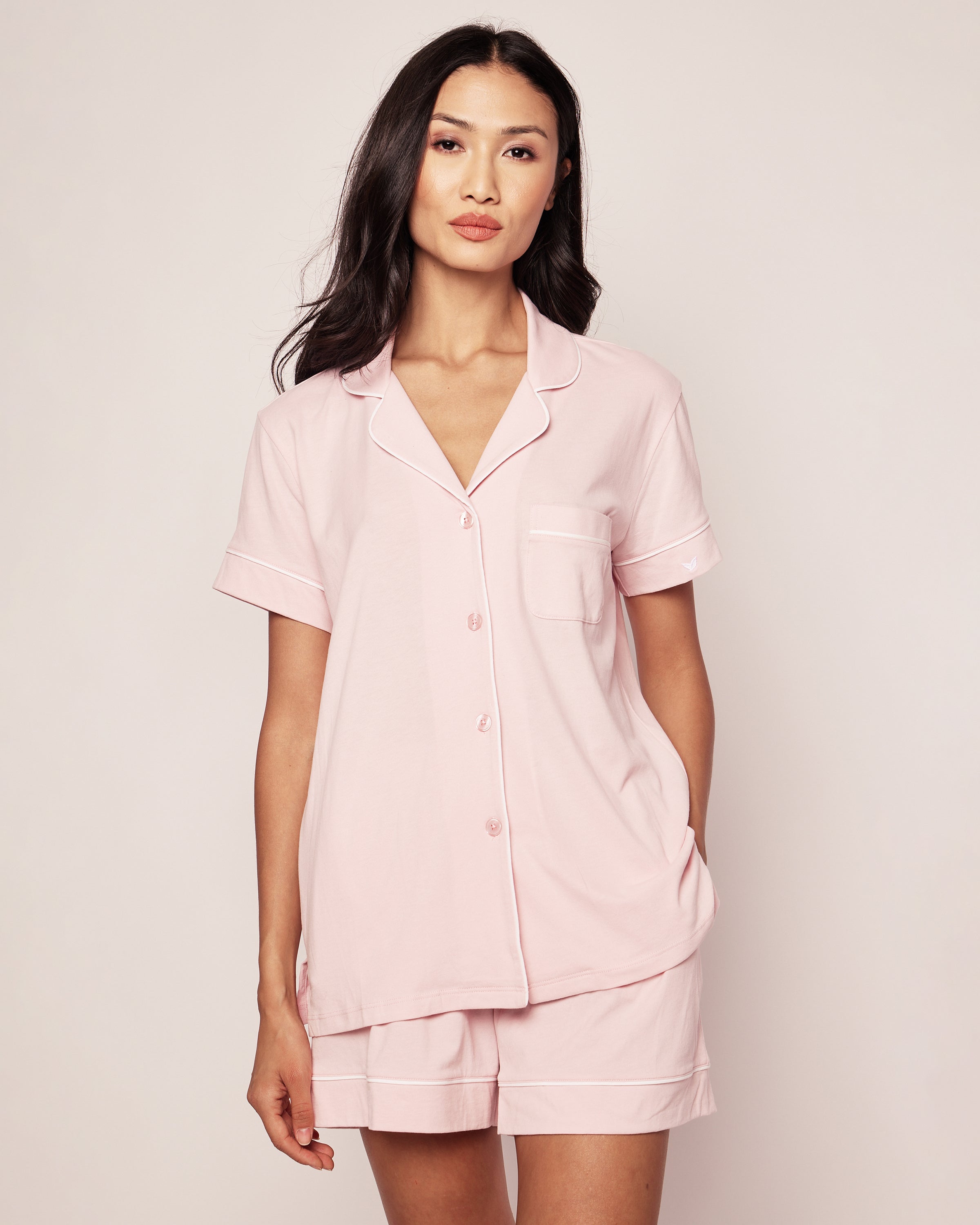 The woman wears a Petite Plume Womens Pima Pajama Short Sleeve Set in Pink, crafted from soft Peruvian Pima cotton. The set includes a short-sleeve button-up top and shorts, elegantly finished with white piping. She stands against a plain background, looking directly at the camera.