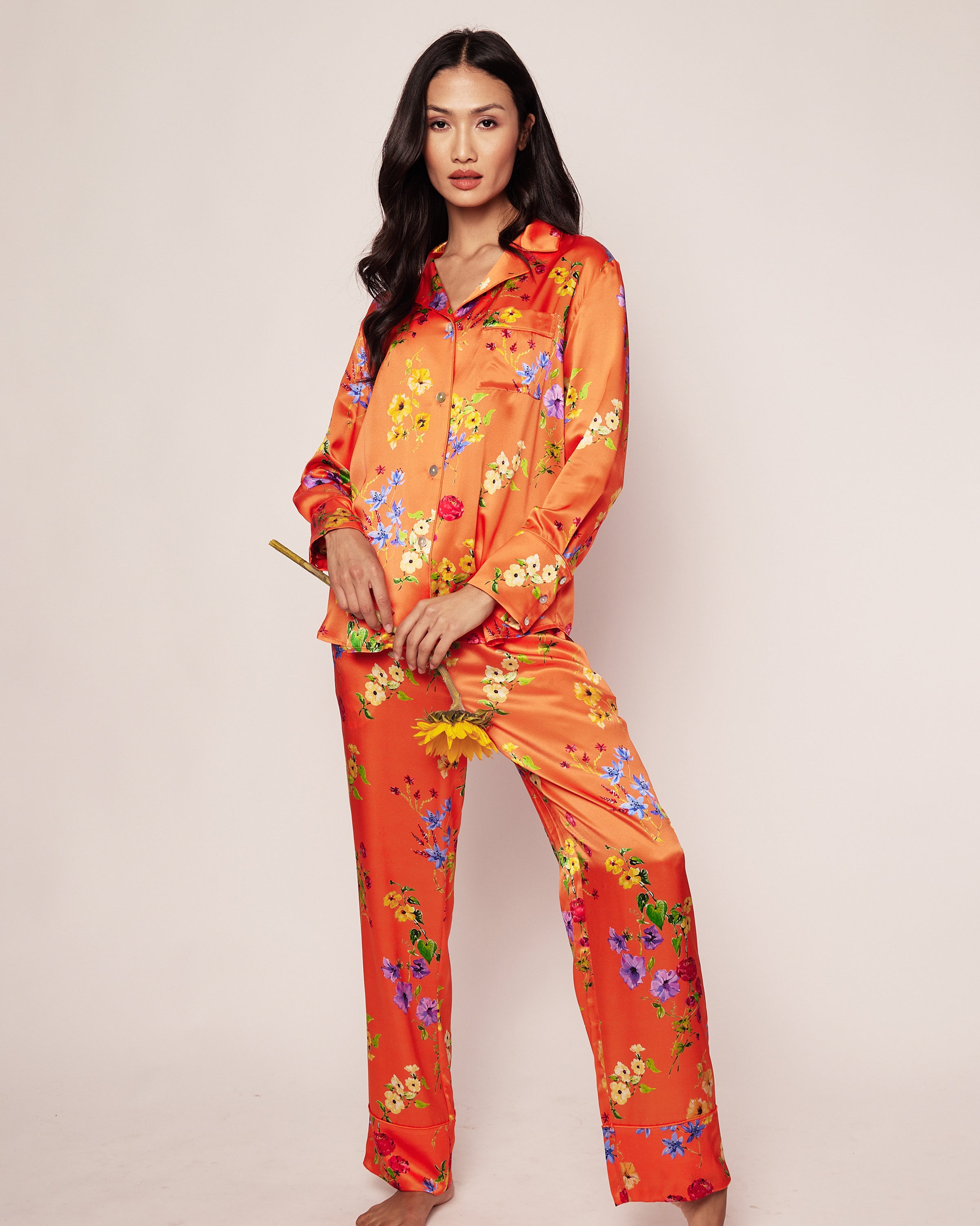 A person confidently poses against a plain background wearing the Womens Silk Pajama Set in Tangerine Brilliant Botanical by Petite Plume, holding a brush with a relaxed expression, embodying luxury.