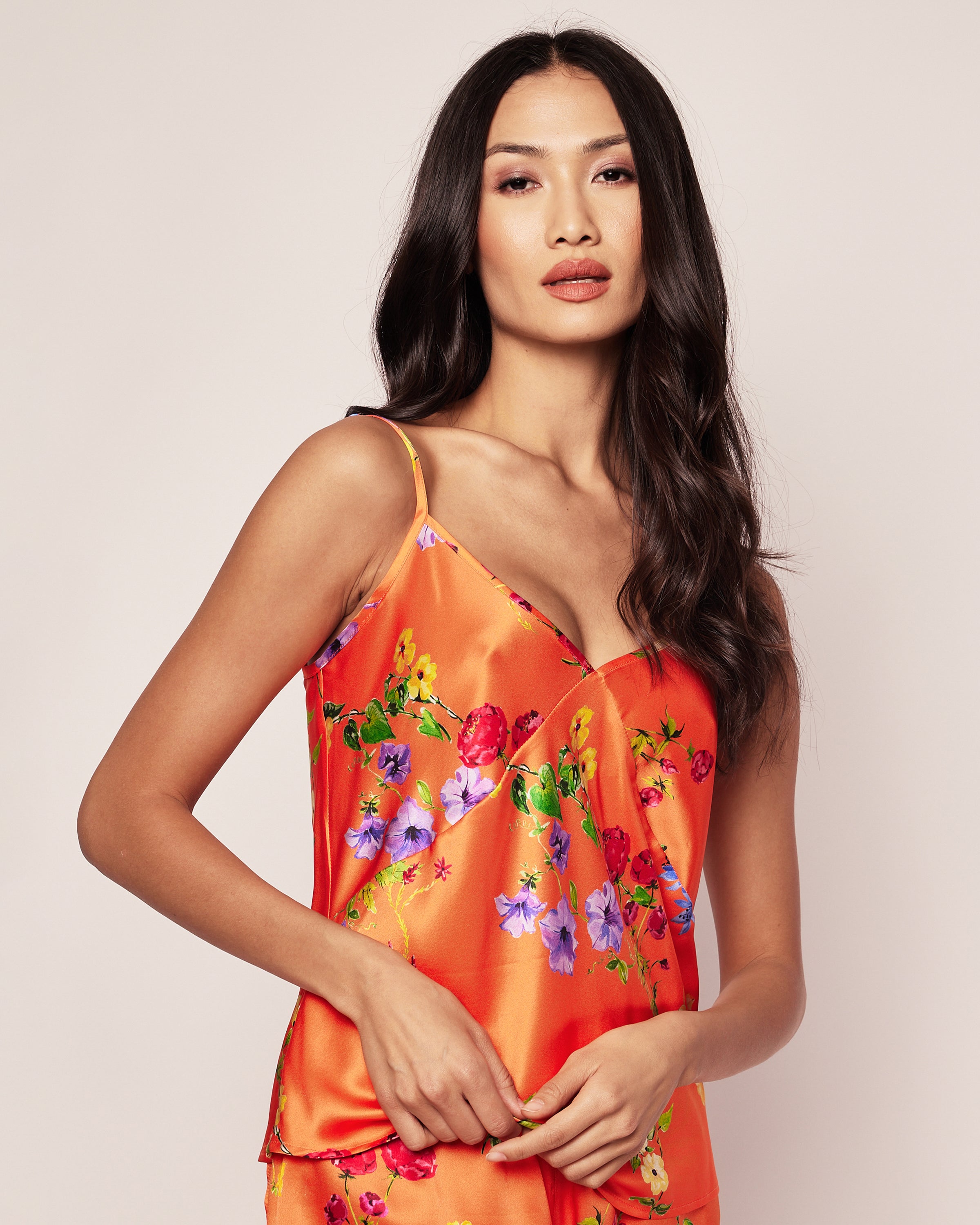 A person with long, dark hair wears the Womens Silk Cami Short Set in Tangerine Brilliant Botanical by Petite Plume. They stand against a plain background, looking toward the camera with a neutral expression.