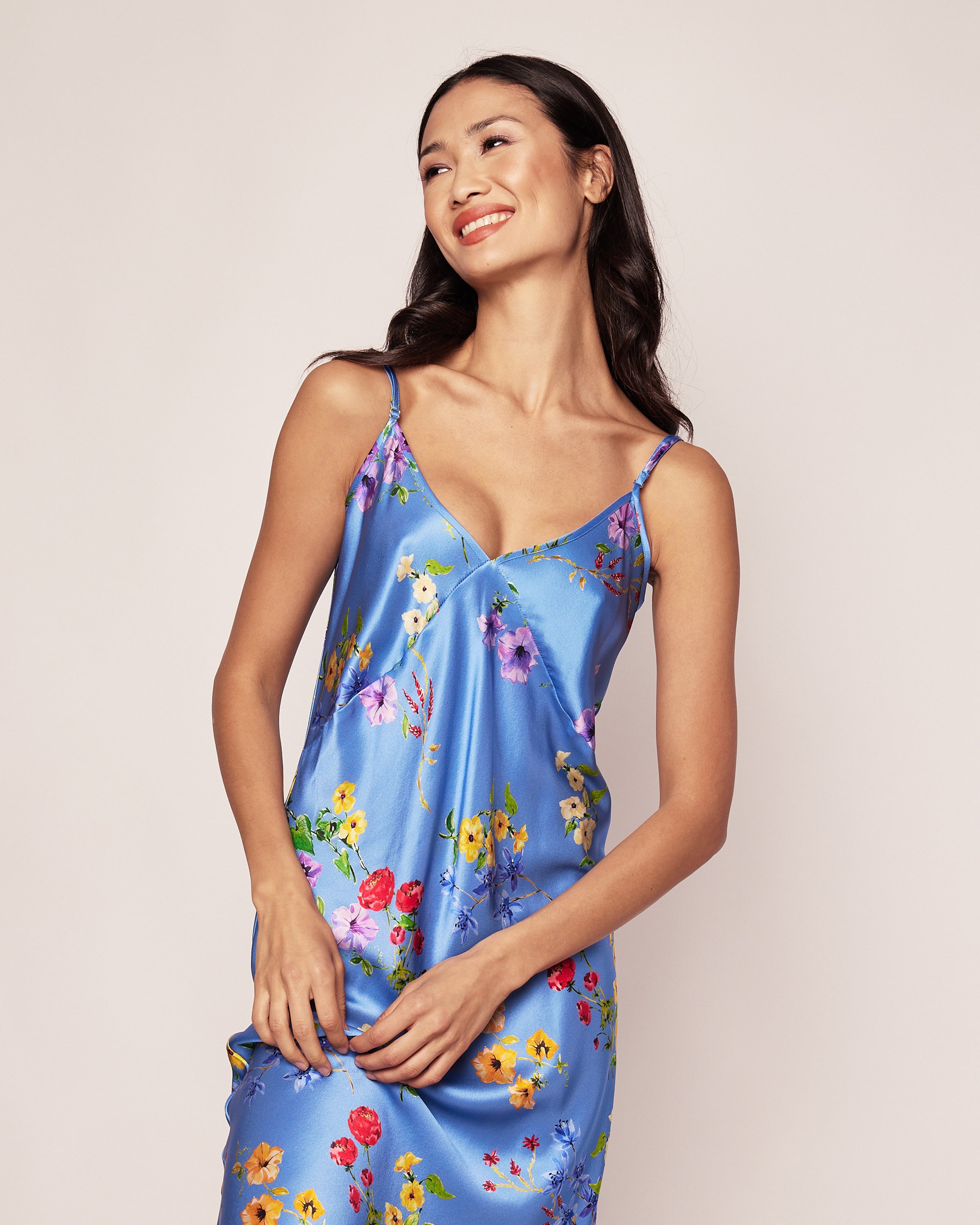 A woman in a Petite Plume Womens Silk Cosette Nightgown in Azure Brilliant Botanical, featuring vibrant botanical flowers, stands smiling against a plain background. Her long dark hair flows over her shoulders as she looks slightly to the side.