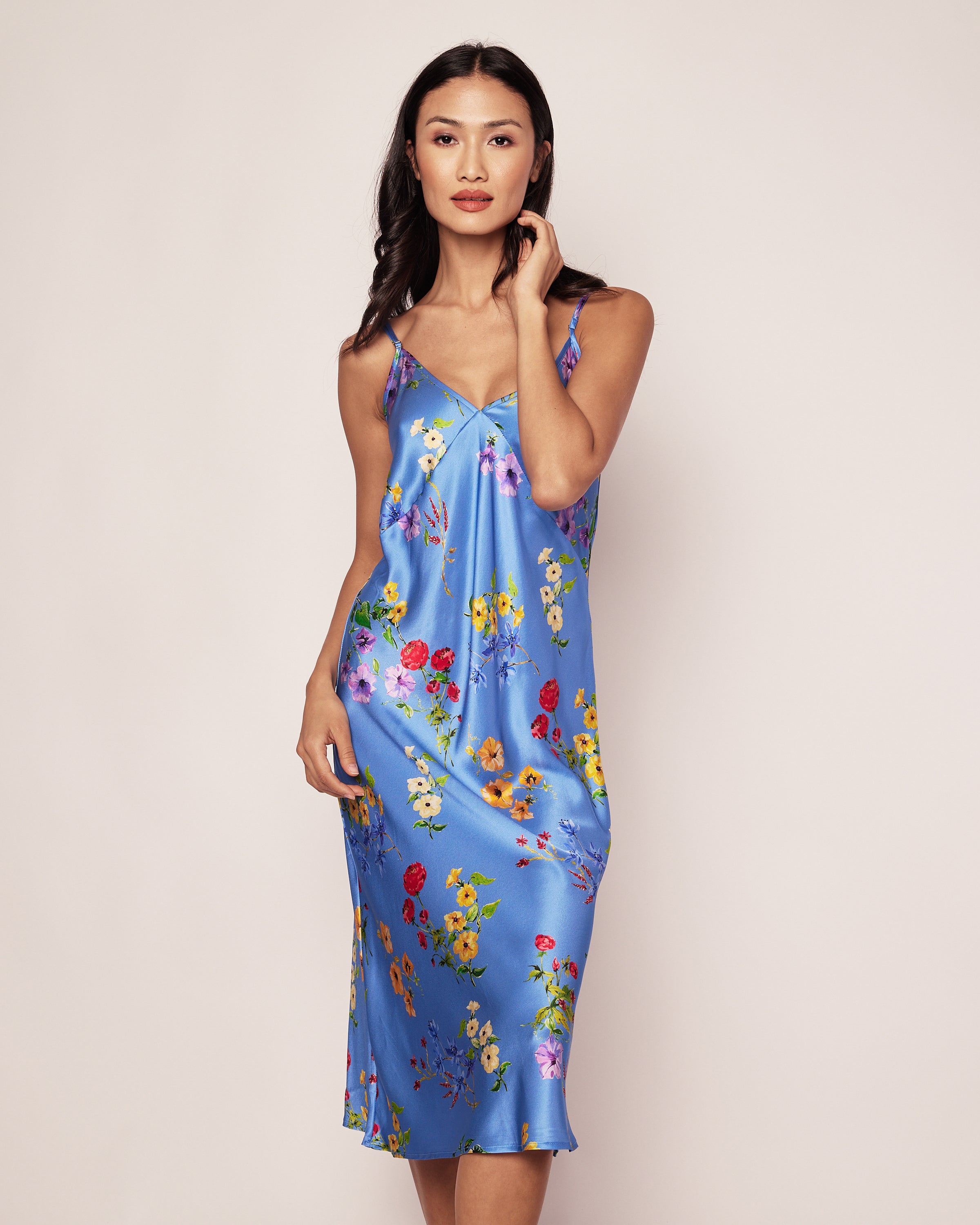 A woman poses against a plain background, wearing the Womens Silk Cosette Nightgown in Azure Brilliant Botanical by Petite Plume. Her hair is down, and she gently touches her face with one hand. The nightgown features thin straps and a relaxed fit reminiscent of luxury sleepwear.