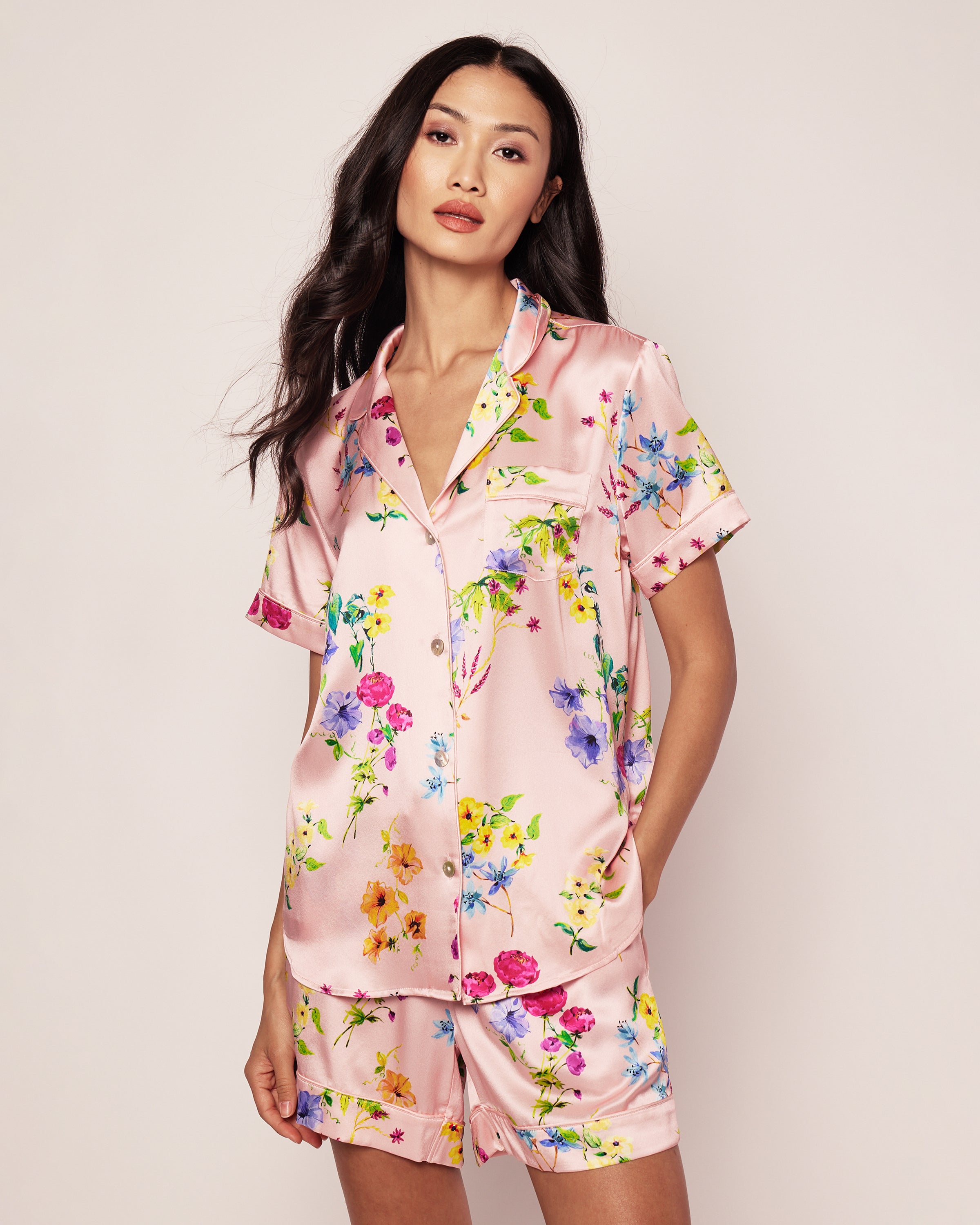 The woman is wearing the Womens Silk Pajama Short Set in Blush Brilliant Botanical by Petite Plume. Her long dark hair cascades as she poses against a plain background. The set features a short-sleeved button-up top and loose-fitting shorts, epitomizing elegance in comfort.