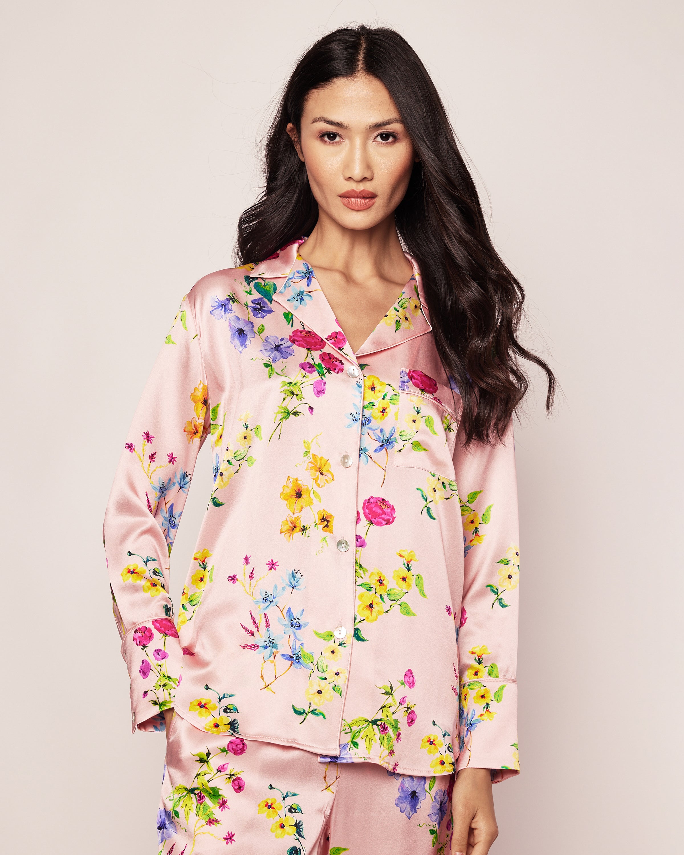 A person with long, dark hair wears the Womens Silk Pajama Set in Blush Brilliant Botanical by Petite Plume. The silk sleepwear features a collared button-up top with a botanical print of yellow, pink, and blue flowers, set against a plain background.