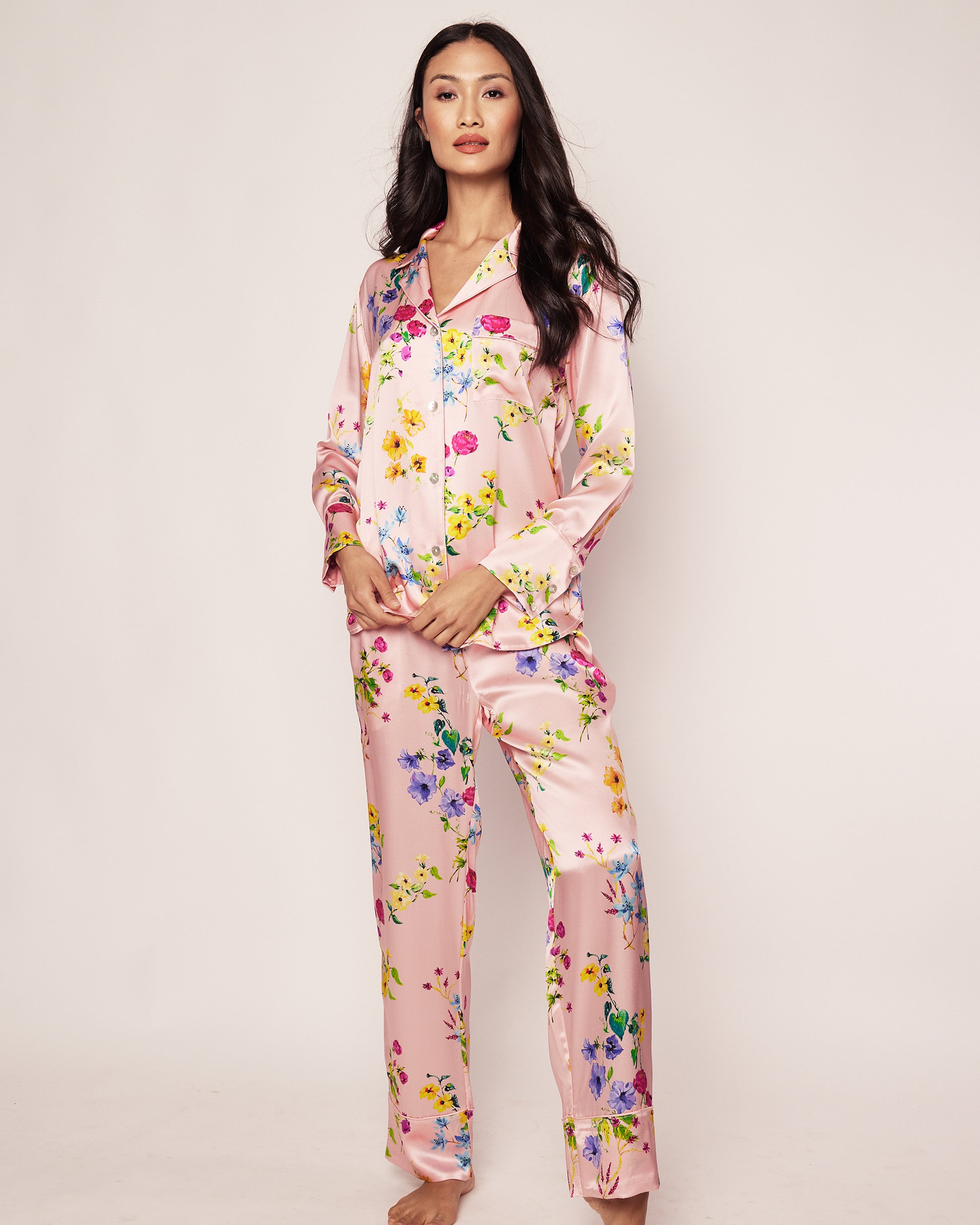 A person stands barefoot against a plain background, wearing the Womens Silk Pajama Set in Blush Brilliant Botanical by Petite Plume. The luxurious pajamas feature long sleeves and a button-up shirt, embodying the elegance of silk sleepwear.