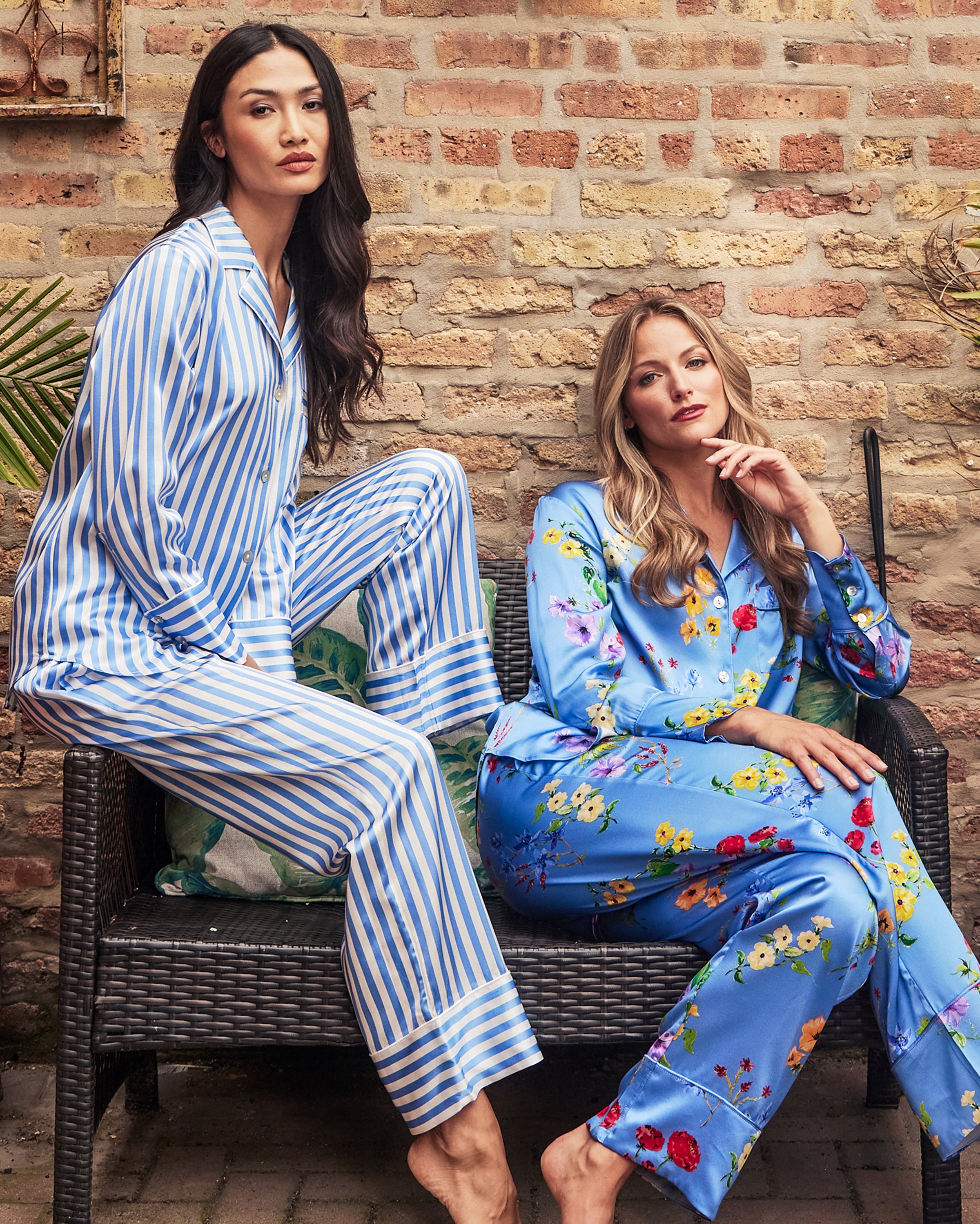 Two women in Petite Plumes luxurious silk pajamas sit on a wicker bench against a brick wall. One flaunts the Womens Silk Pajama Set in Azure Stripe, while the other showcases a blue floral pattern, exuding relaxed yet fashionable vibes in a cozy outdoor setting.