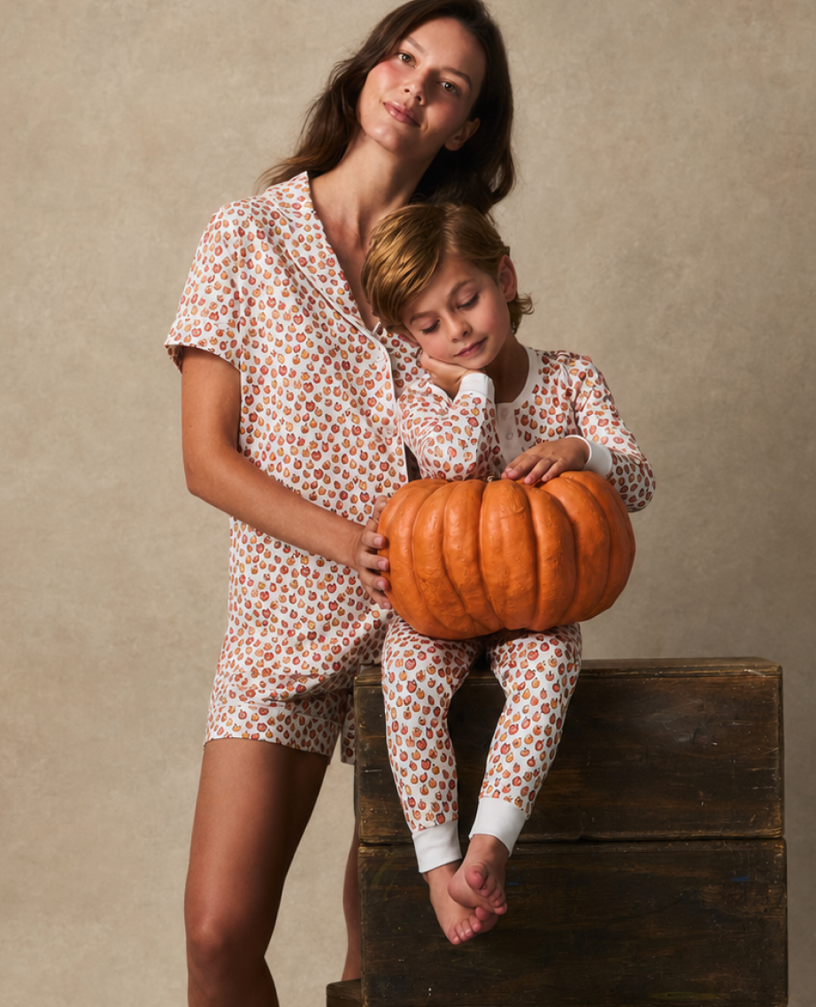 A woman and a child in matching patterned pajamas pose together; the child holds a large pumpkin and rests their head on the womans shoulder while sitting on a wooden box against a neutral background.