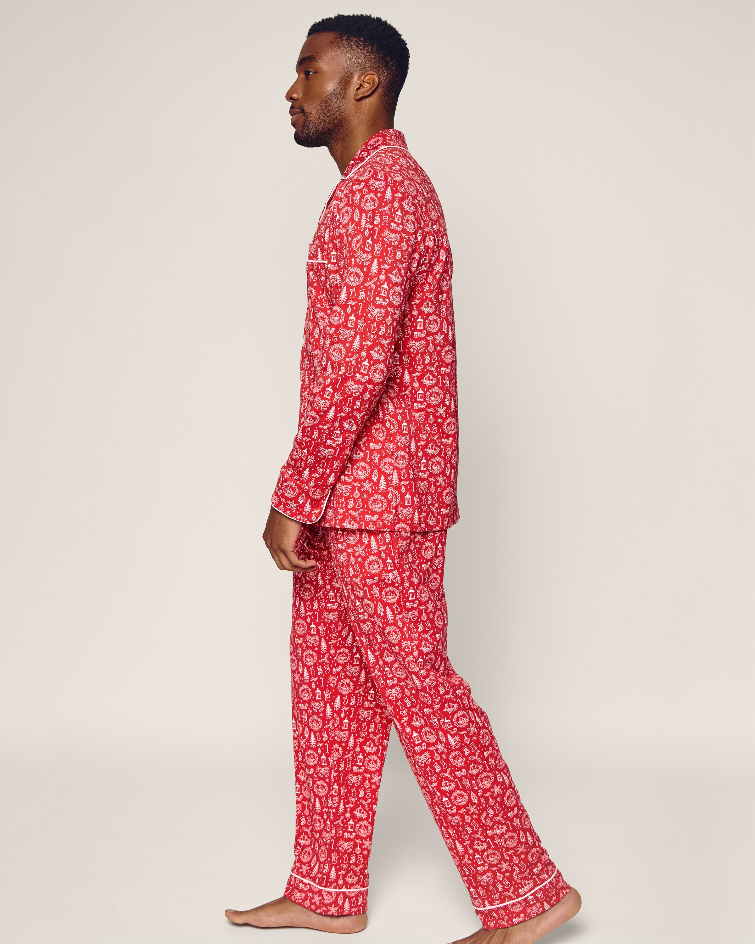 A person stands barefoot in profile wearing the Petite Plume Mens Pima Pajama Set, featuring the Whimsical Winter Saks x Petite Plume design. The luxurious Peruvian Pima cotton set includes a red long-sleeved shirt and pants with a white floral pattern against a plain, neutral background.