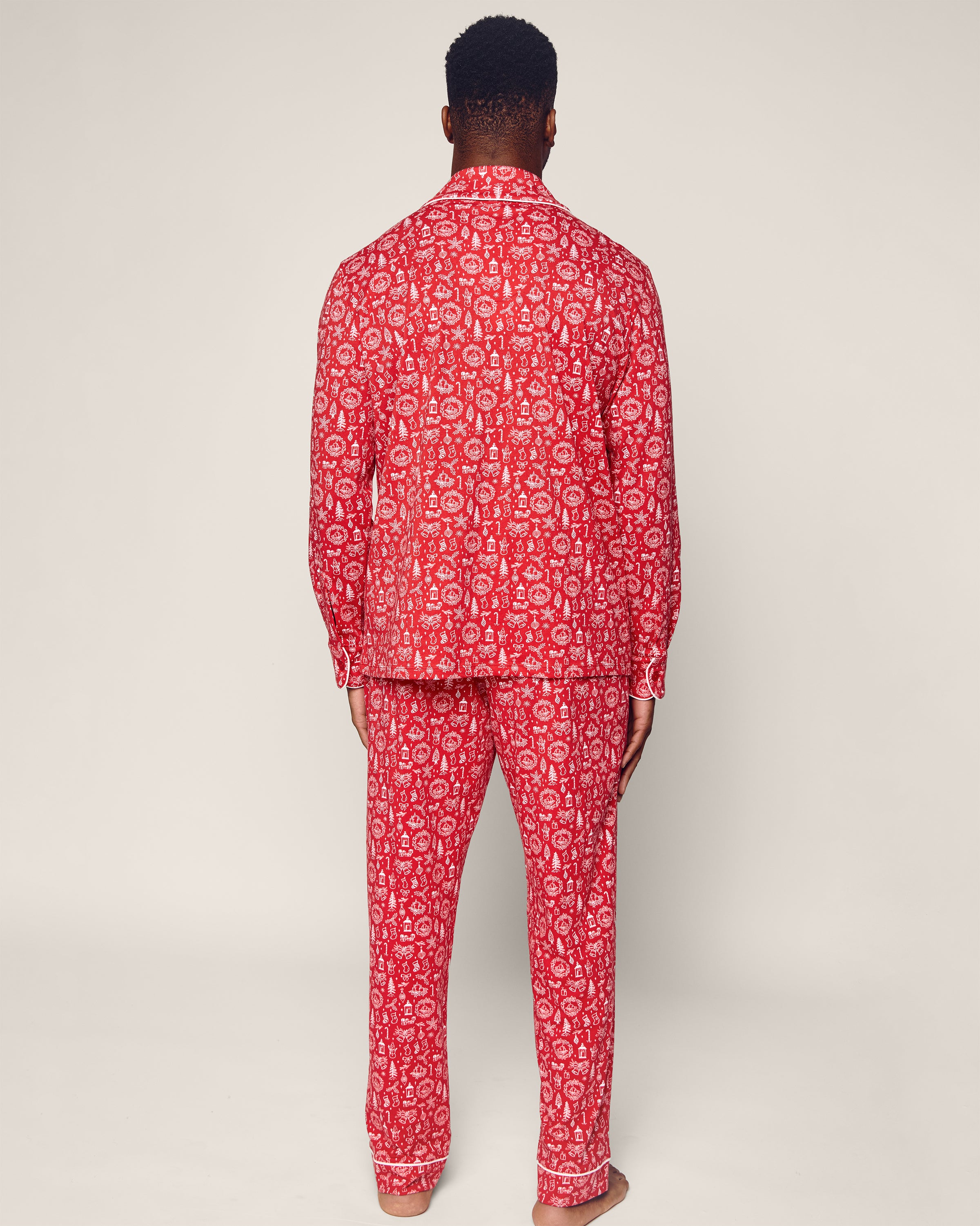 A person stands barefoot against a plain background, wearing Petite Plumes Men’s Pima Pajama Set in Whimsical Winter Saks. The luxurious red sleepwear, made of Peruvian Pima cotton, features a white floral pattern and includes long-sleeve shirt and pants.