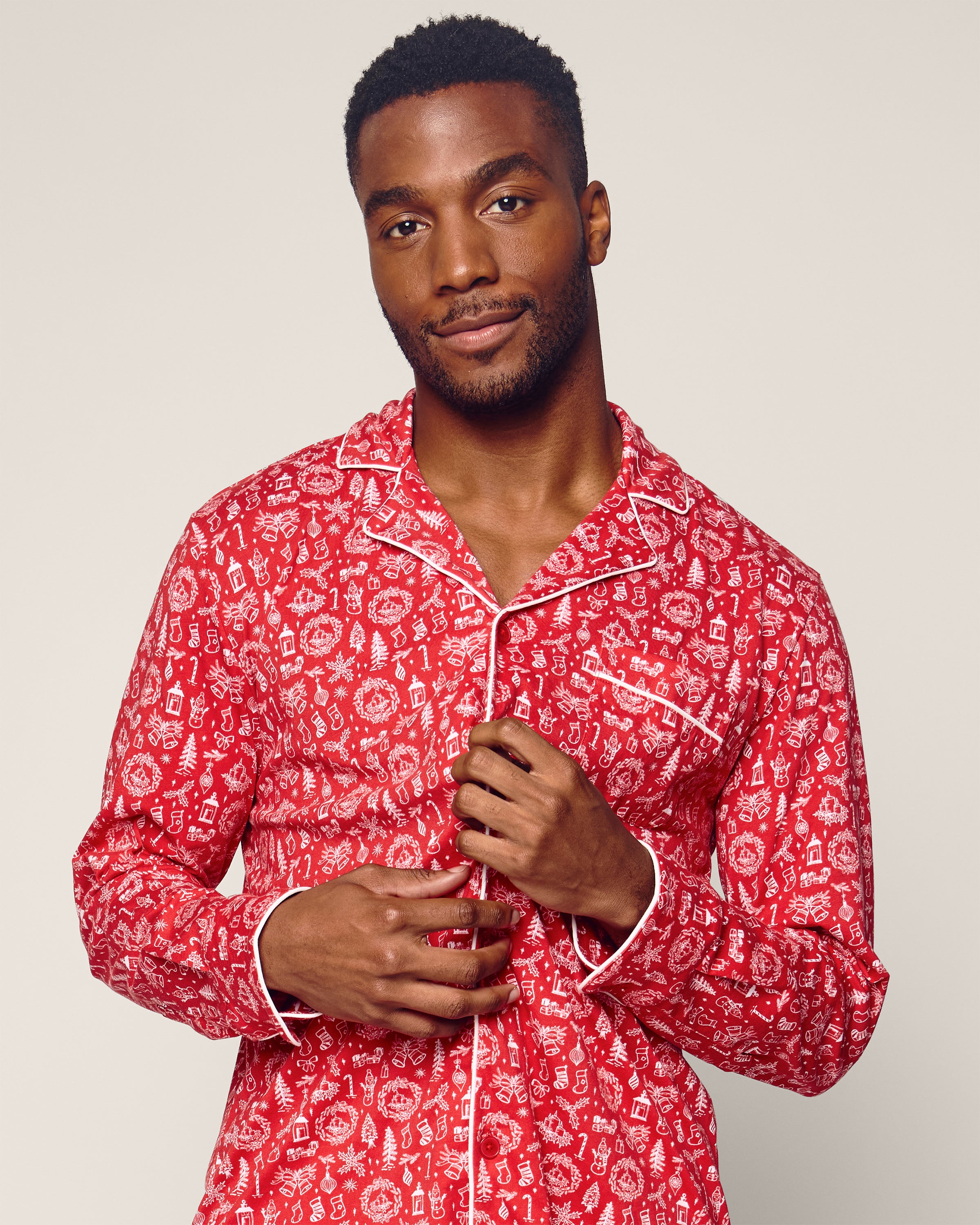 A person wearing Petite Plumes Mens Pima Pajama Set in Whimsical Winter, featuring a red pattern and white piping, holds the lapels while standing against a neutral background. They have short hair, looking at the camera with a subtle smile.