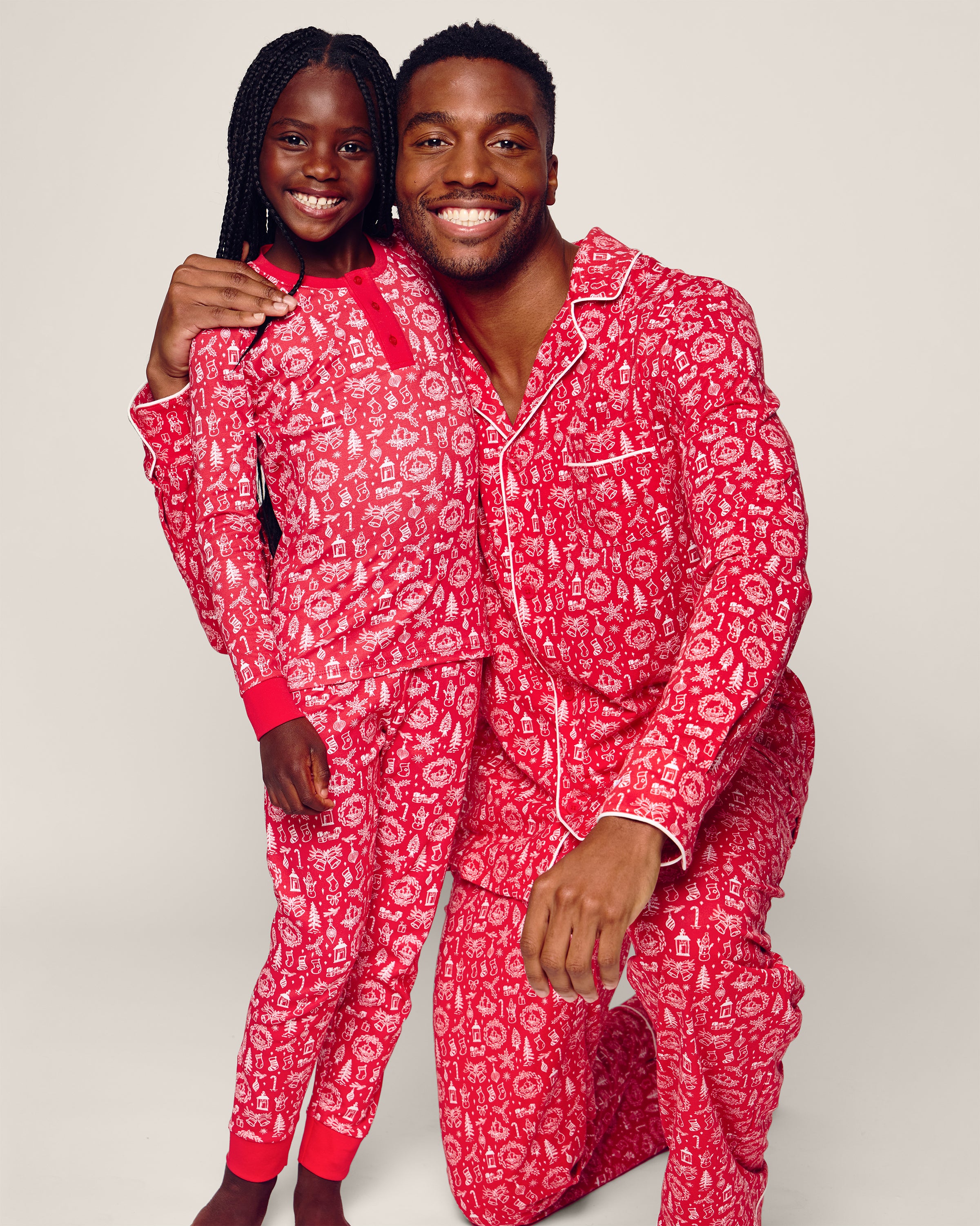 A man kneels beside a girl, both smiling in matching red pajamas made from luxurious Peruvian Pima cotton by Petite Plume. The background is plain and light-colored.