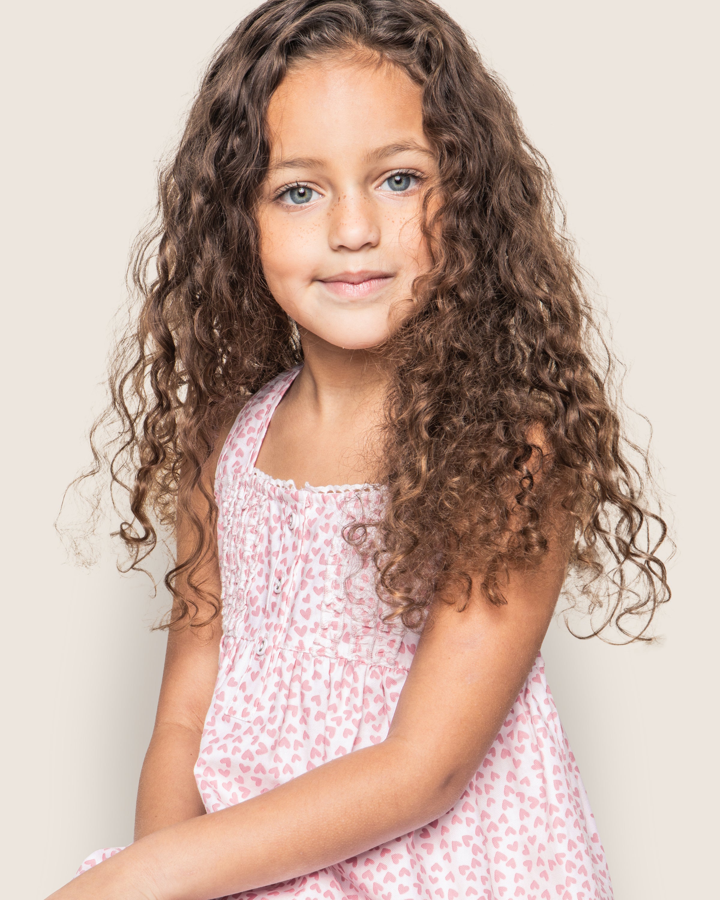 The young girl with long, curly brown hair smiles softly at the camera, wearing Petite Plumes Girls Twill Charlotte Short Set in Sweethearts. Her innocent charm stands out against the plain background, capturing the luxurious elegance of high-end childrens fashion.