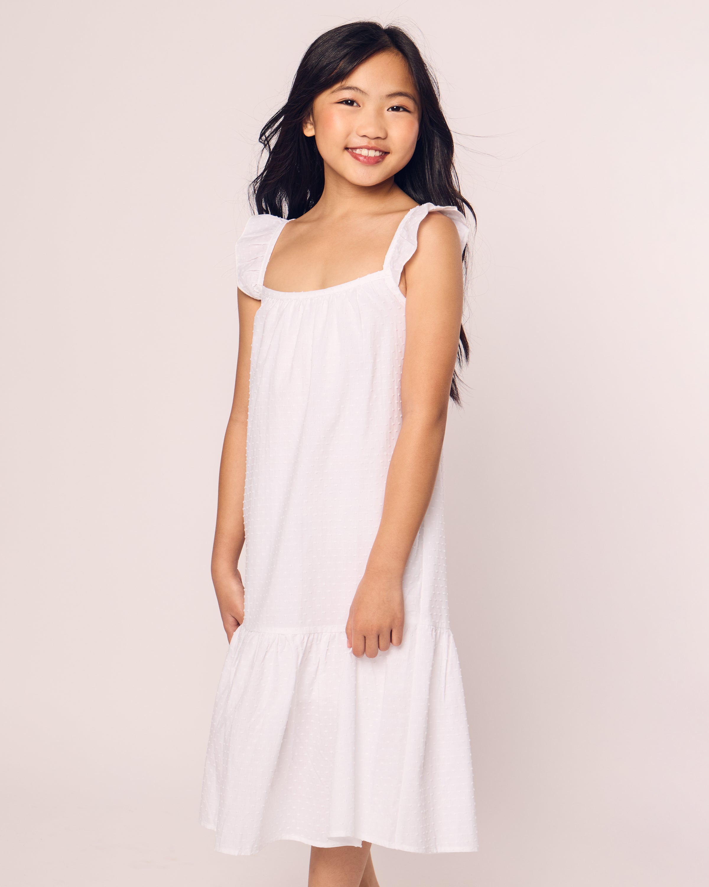 A young girl smiles in a Petite Plume Girls Swiss Dots Celeste Day Dress, a white sleeveless cotton outfit with ruffled shoulders and subtle texture ideal for luxury wear. Her long hair flows over her shoulders against a plain, light-colored background.