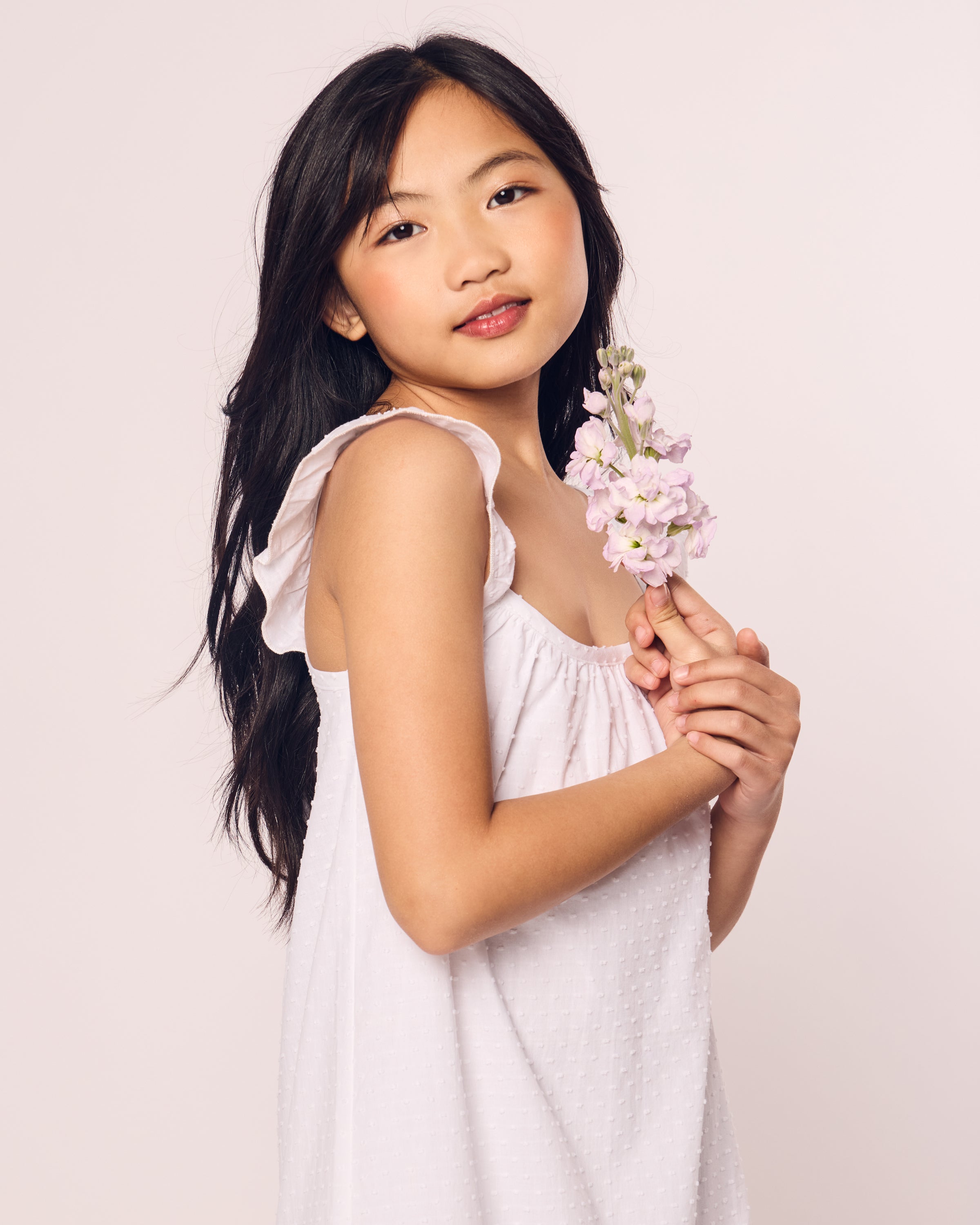 A girl with long dark hair wears Petite Plumes Girls Swiss Dots Celeste Day Dress in White. She holds a small bouquet of purple flowers and gazes gently at the camera against a plain, light background.