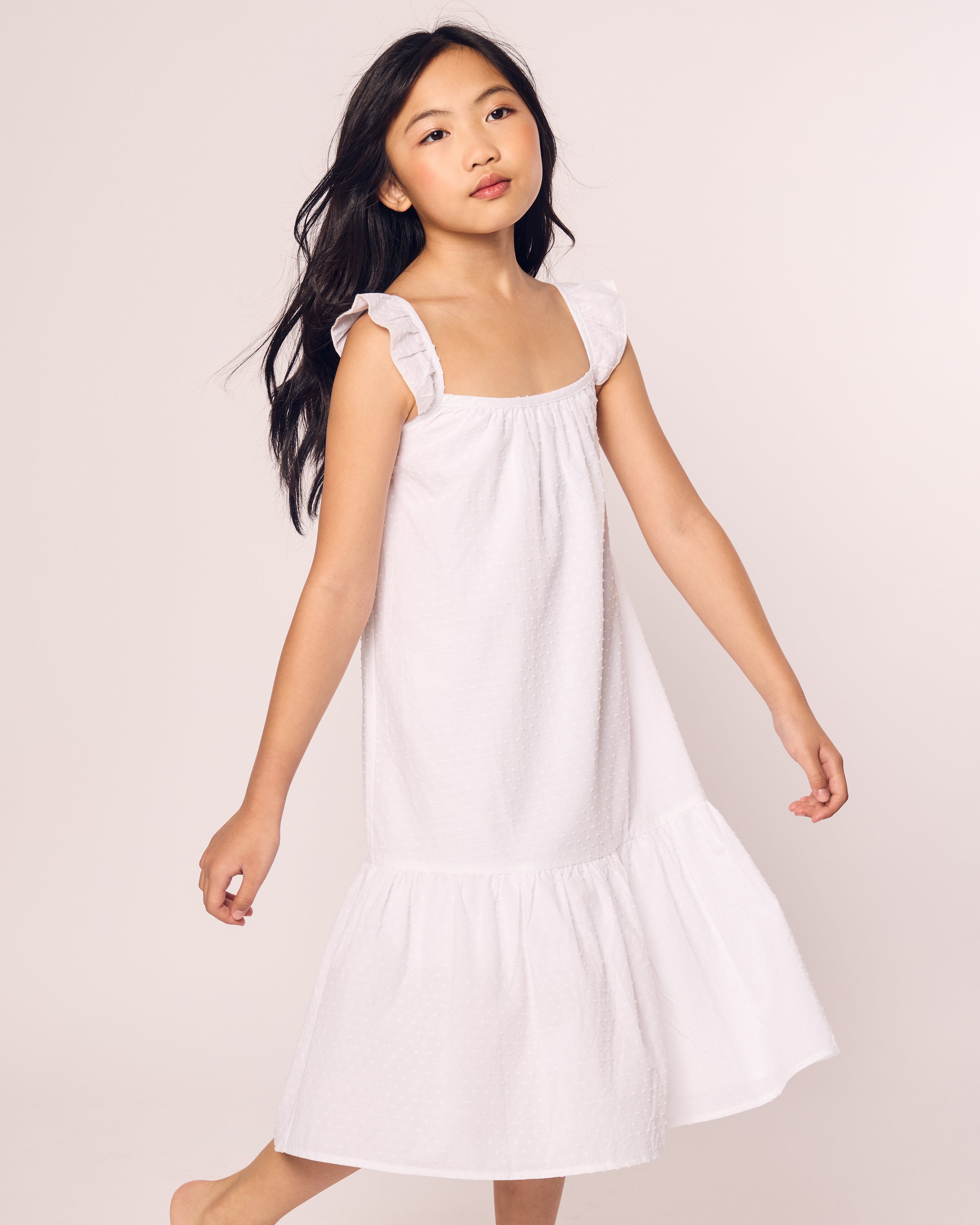 A young girl with long, dark hair wears the Petite Plume Girls Swiss Dots Celeste Day Dress in White. This sleeveless dress, featuring ruffled straps and flowing to her calves, highlights her side profile and slight smile against a plain, light-colored background.