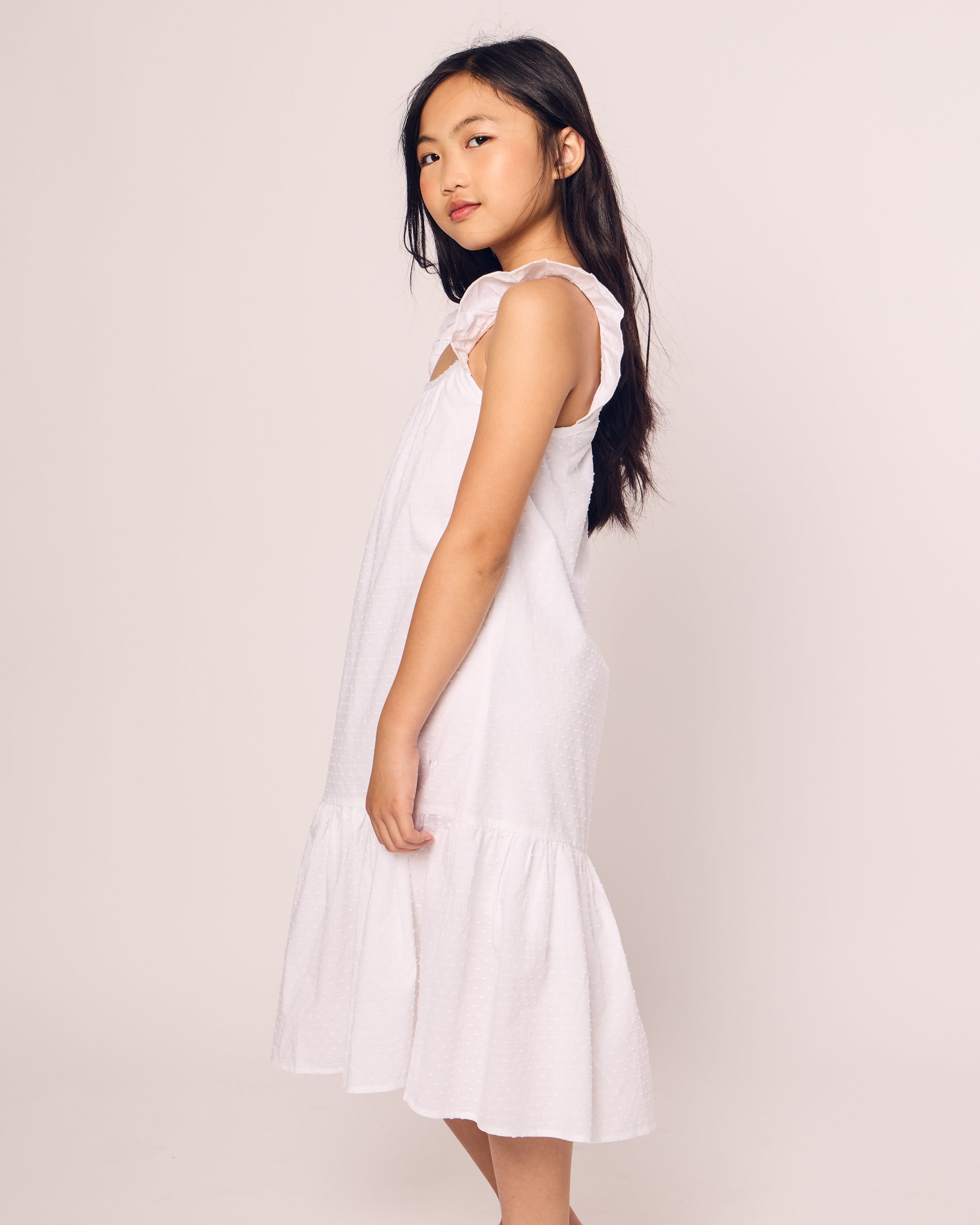 A young girl with long dark hair stands sideways, looking at the camera. She wears the Petite Plume Girls Swiss Dots Celeste Day Dress in White, featuring ruffled shoulders and a flared hem. The background is a simple off-white.