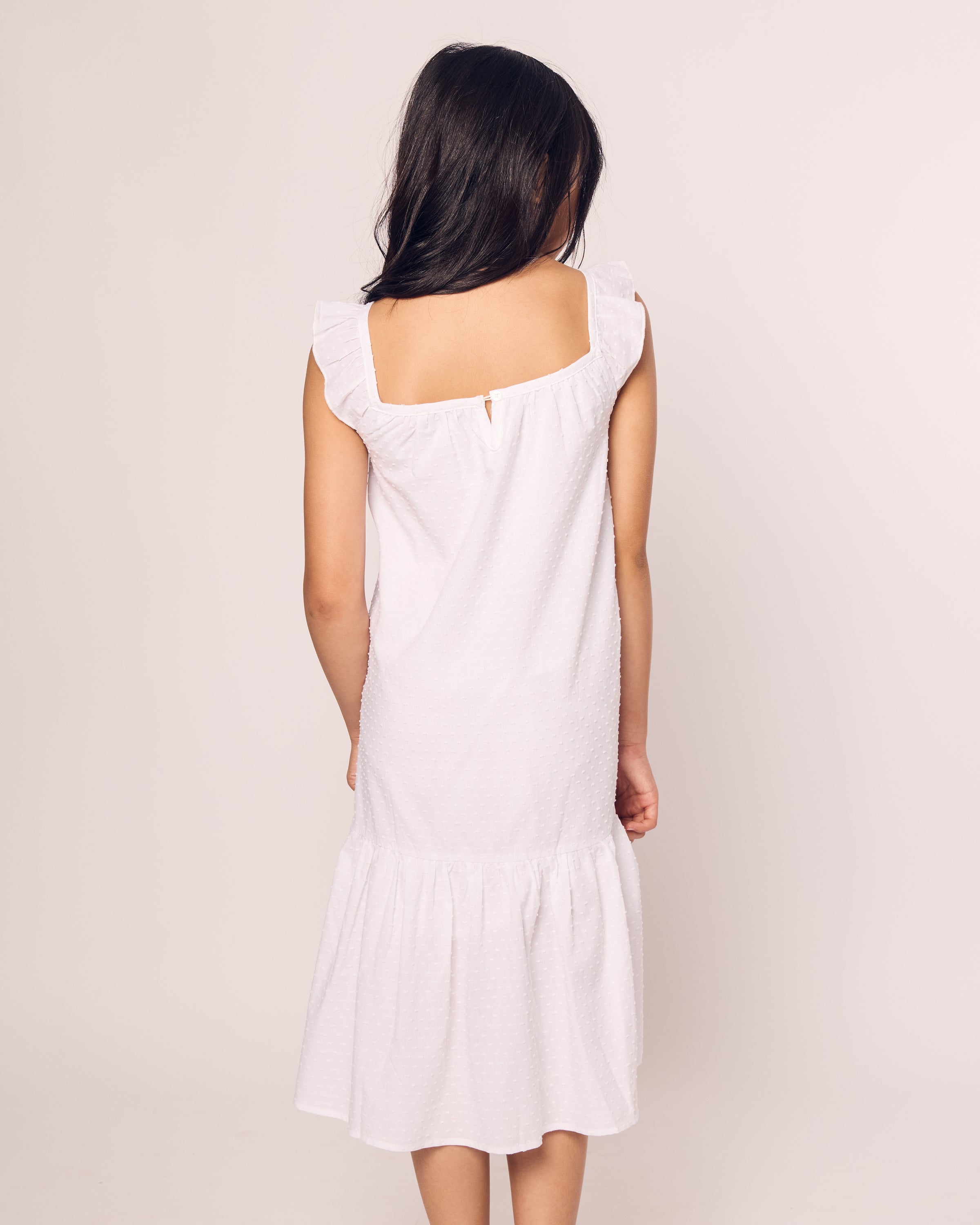 A person with long dark hair is shown from the back wearing Petite Plumes Girls Swiss Dots Celeste Day Dress in white, featuring ruffled shoulders and a knee-length hem, standing against a plain, light-colored background.