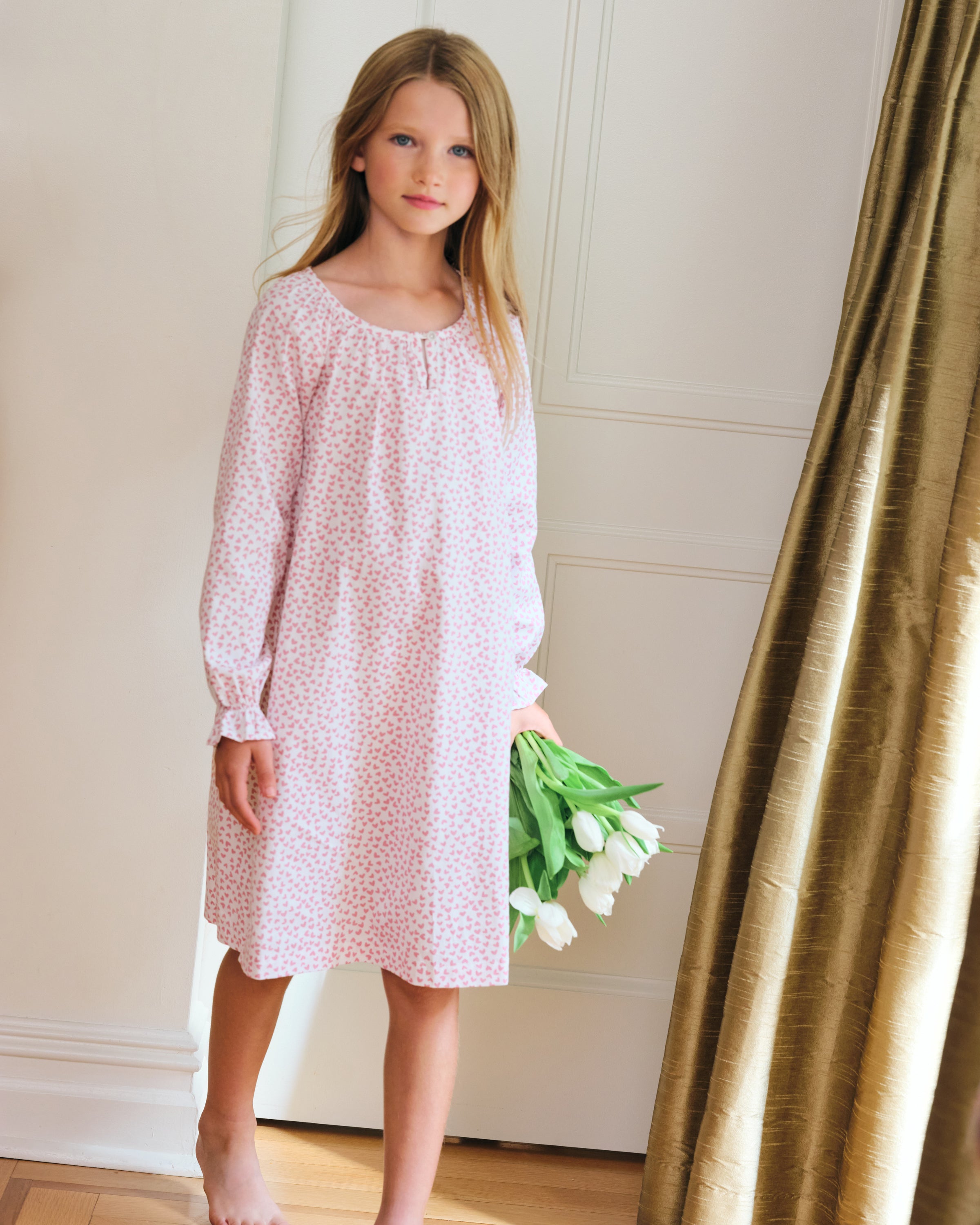 The Girls Twill Delphine Nightgown in Sweethearts by Petite Plume features long sleeves, white fabric with a pink floral pattern, is made from yarn-dyed cotton, and has a ruffled neckline with a small bow and ruffled sleeve cuffs.