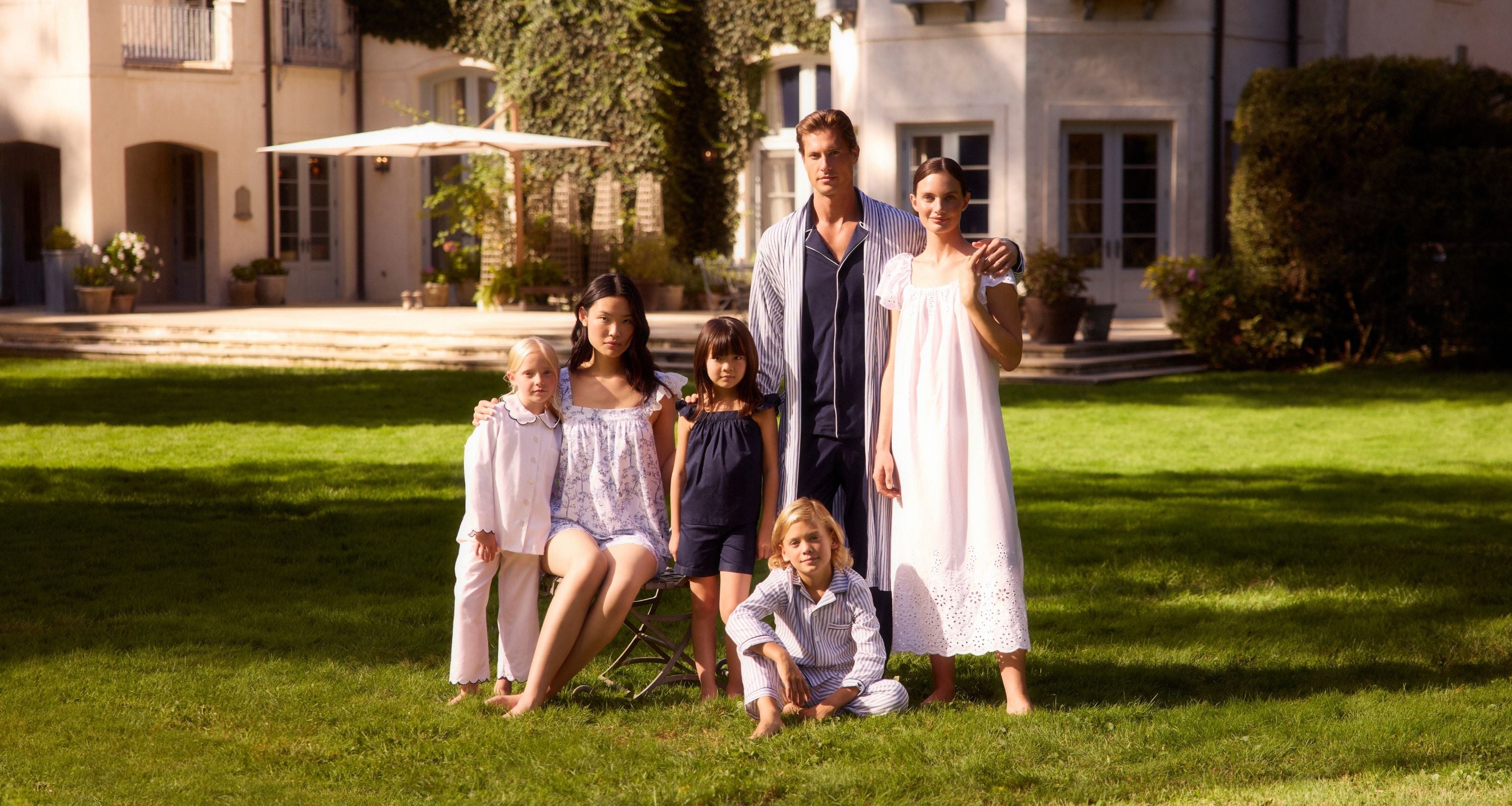 Six people, including four children and two adults, pose together on a sunny lawn in front of a large house, dressed in light, summery clothes. Some are standing, others are seated, all facing the camera.