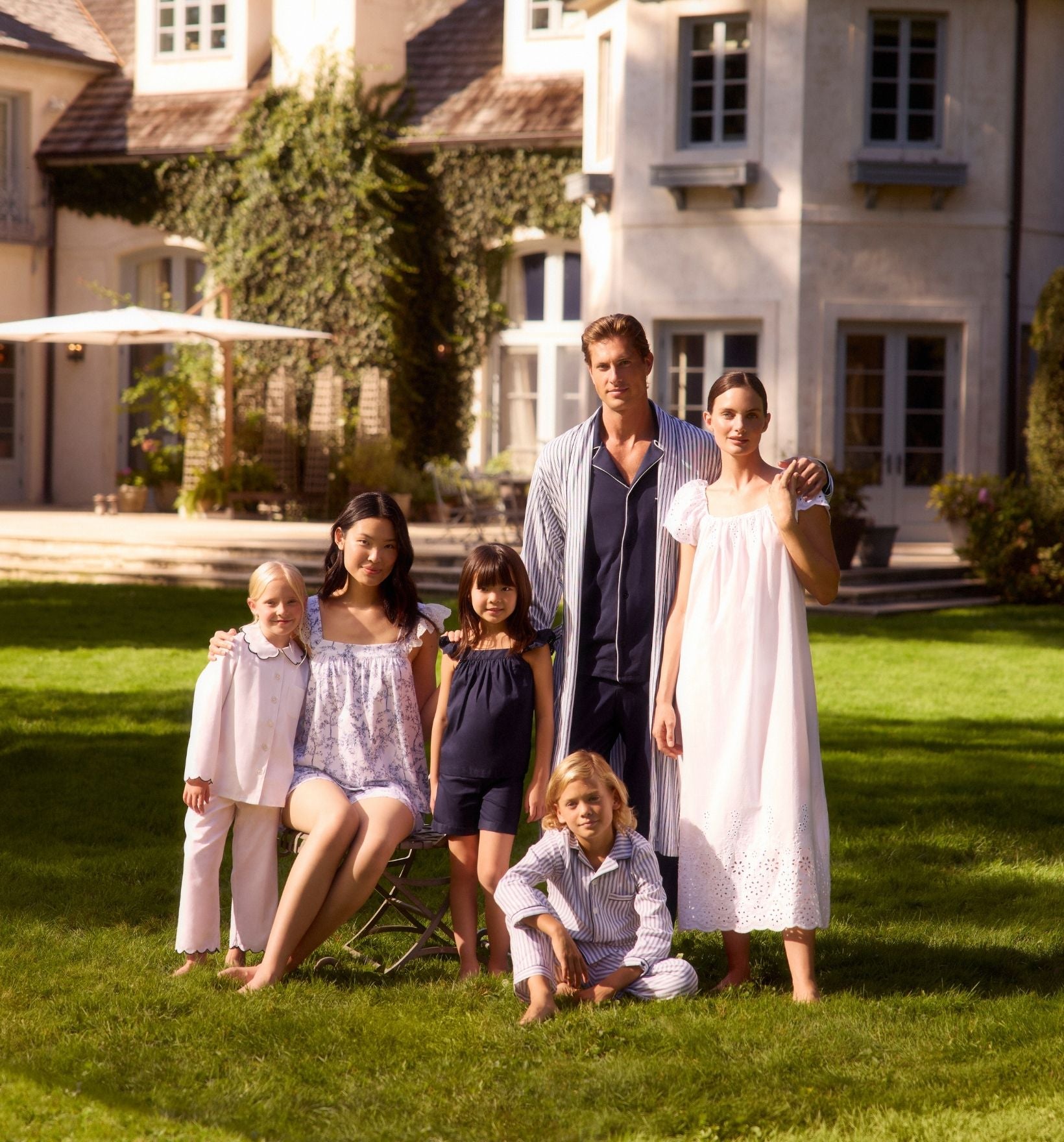 A family of six, wearing light-colored pajamas, poses together on a green lawn in front of a large house with windows, ivy, and a sunlit patio.