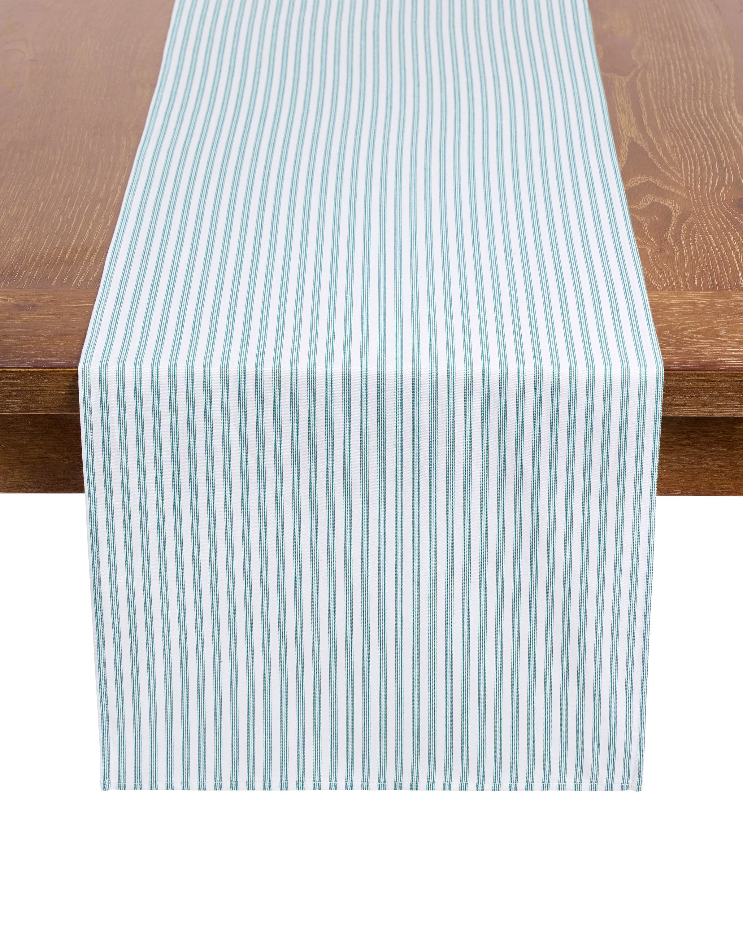 A wooden table boasts the Petite Plume Signature Twill Table Runner in Emerald Ticking, featuring white and teal vertical stripes. Draped down the center, this cotton runner adds a simple yet elegant touch to any festive tablescape.