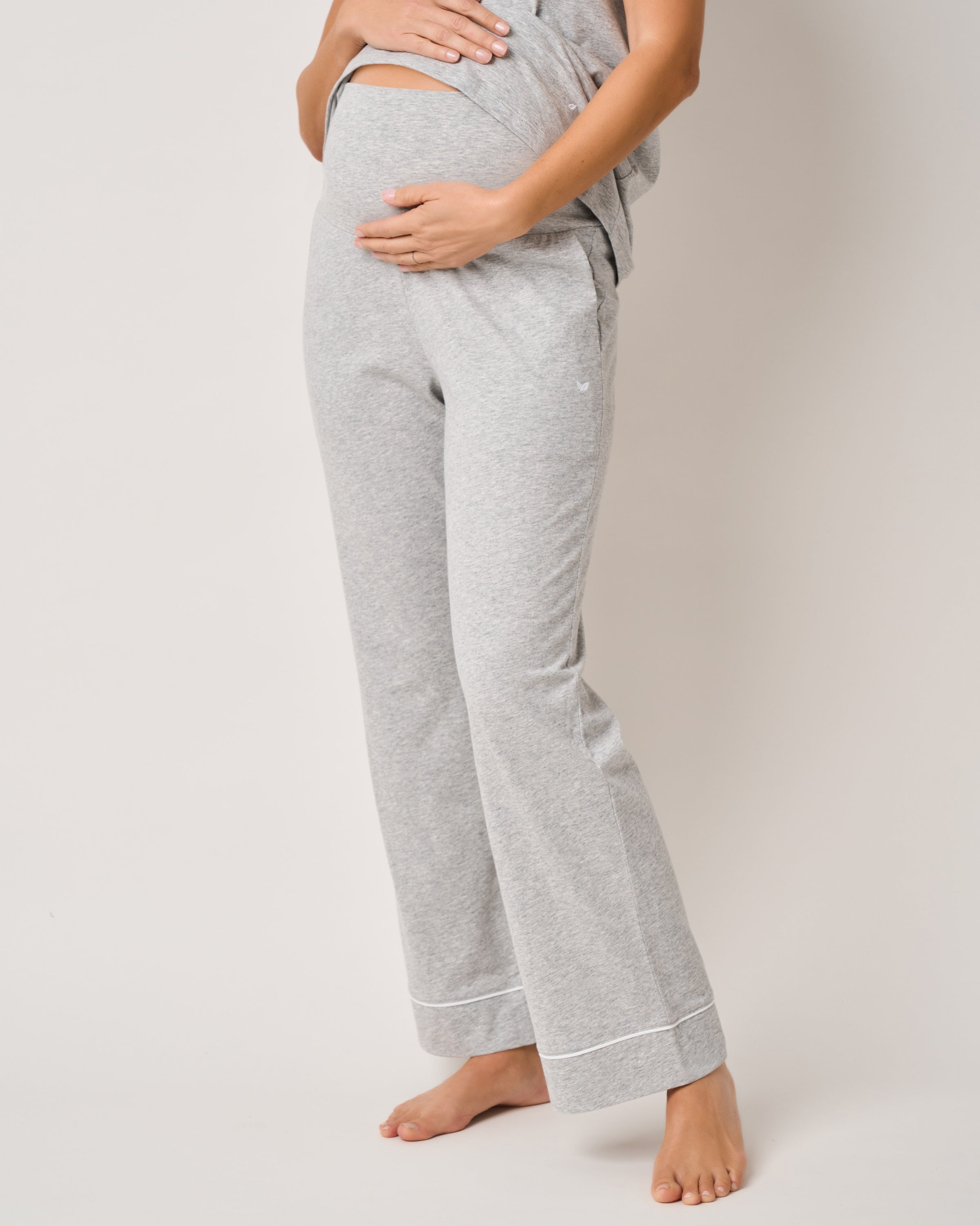 A person is wearing Petite Plumes Womens Pima Maternity Pants in Light Heather Grey, cradling their midsection for pregnancy comfort. The loose-fitting top and long pants embrace maternity ease. They are barefoot against a plain white background.