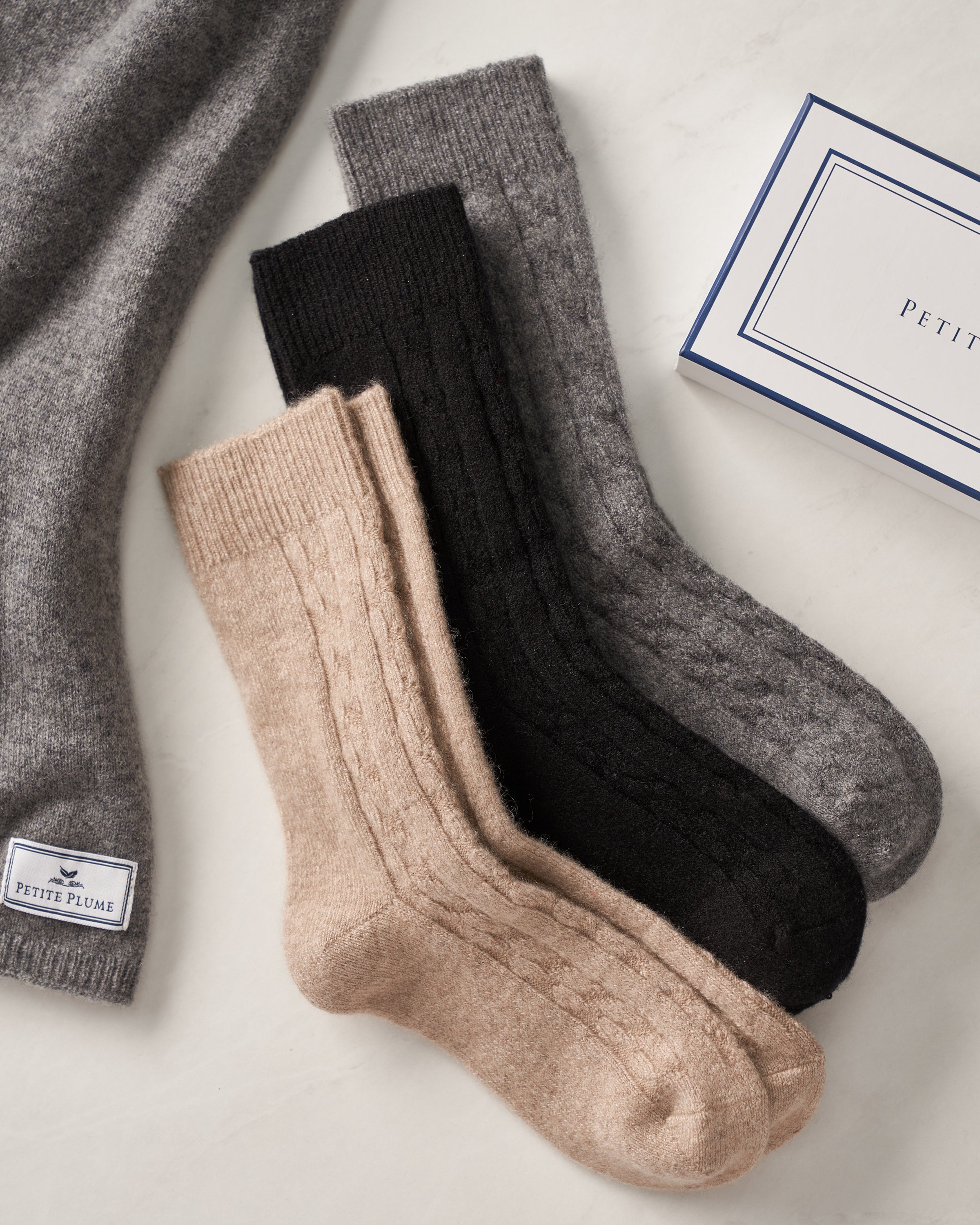 Three pairs of womens cashmere socks in beige, black, and gray reveal their plush texture beside a Petite Plume box. A hint of a luxurious comfort gray garment with the same brand label peeks from the top left corner.