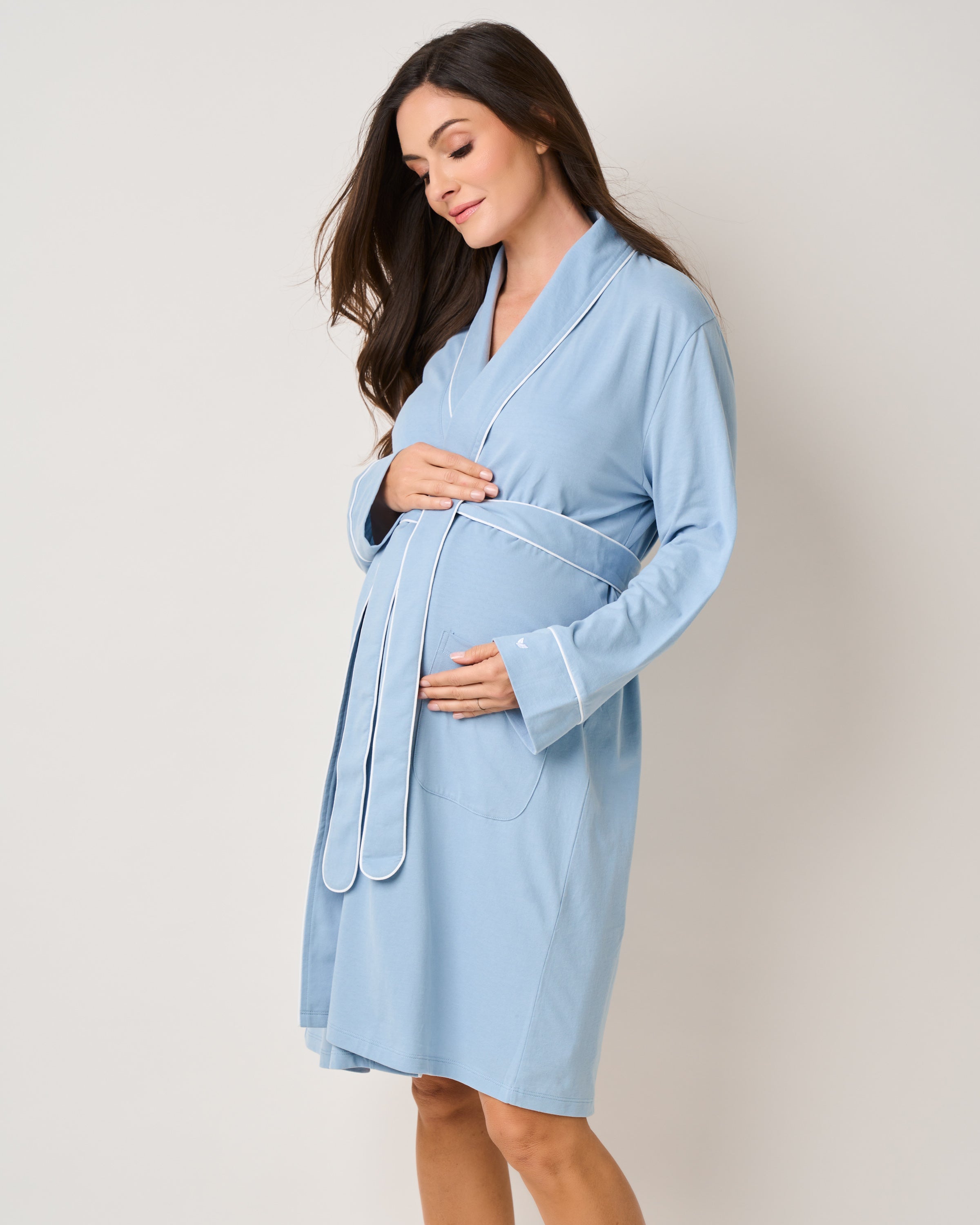A pregnant woman in Petite Plumes The Must Have Maternity Set in Periwinkle cradles her bump in a lightweight, breathable robe. Her long brown hair frames a serene expression as she stands against a plain light gray backdrop.